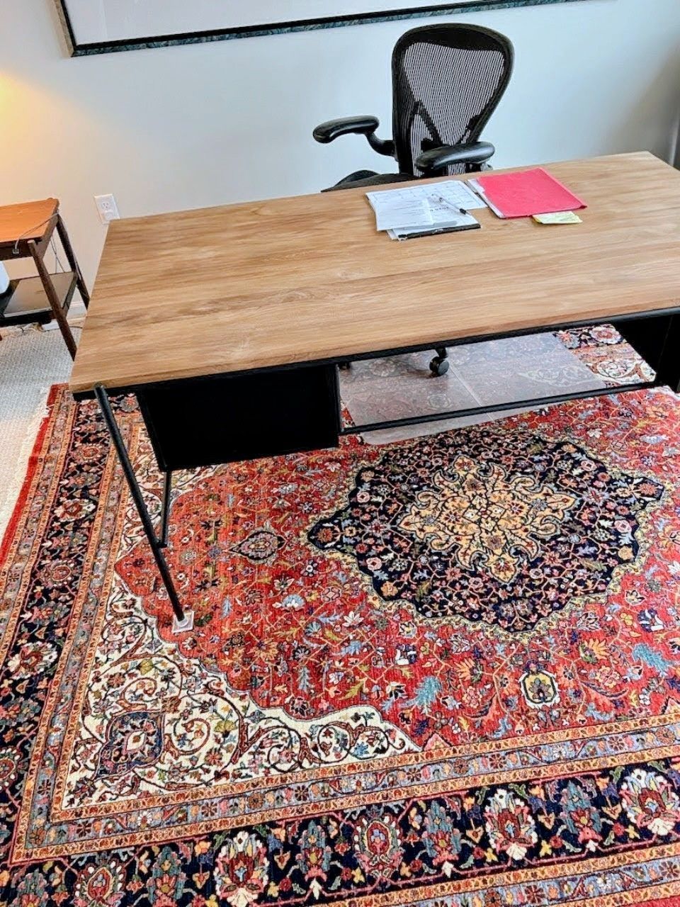 Office with wooden desk, black chair, and ornate red rug. Papers and folder on the desk.
