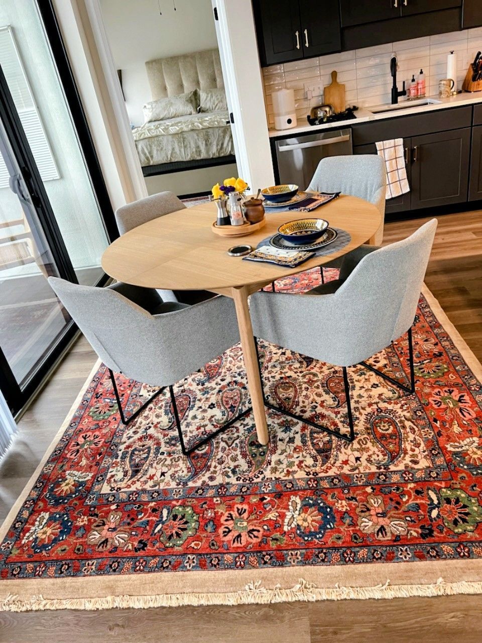 Dining area with wooden table, four gray chairs, and patterned red rug. Kitchen and bedroom in background.