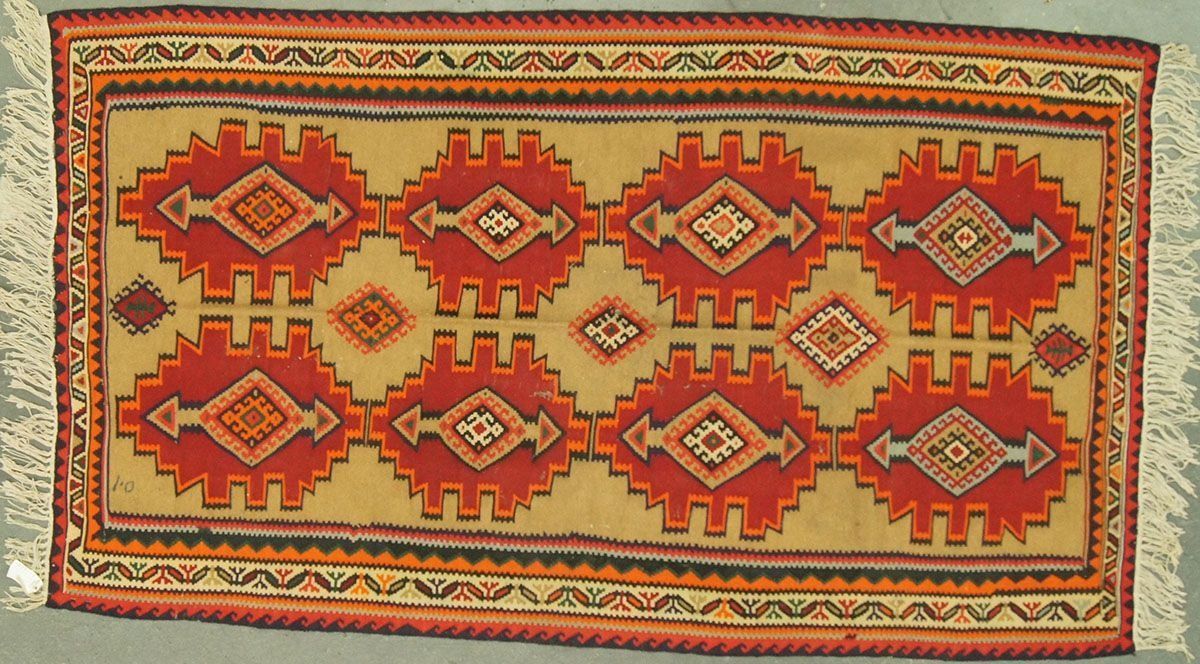 Rectangular rug with eight red, diamond-shaped motifs on a tan background, bordered by decorative bands.