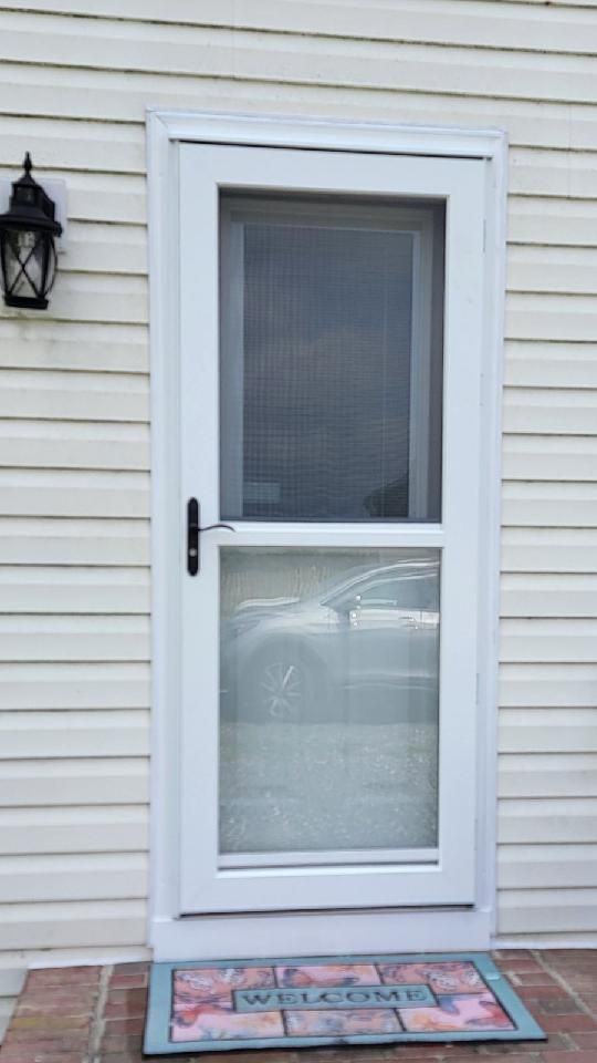 A white screen door is sitting on a brick porch next to a welcome mat.