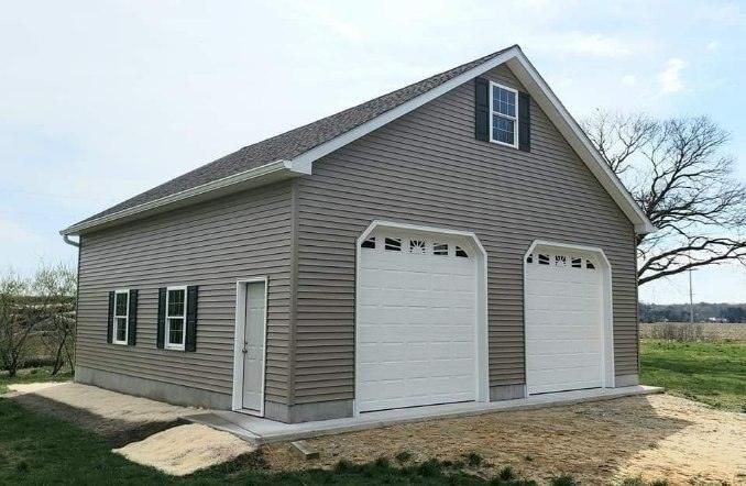 A garage with two garage doors and a roof is sitting in the middle of a grassy field.