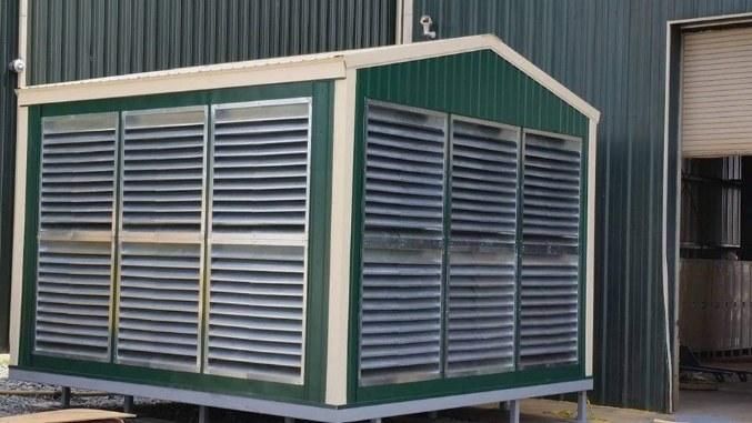 A green shed with shutters on the side is sitting in front of a building.