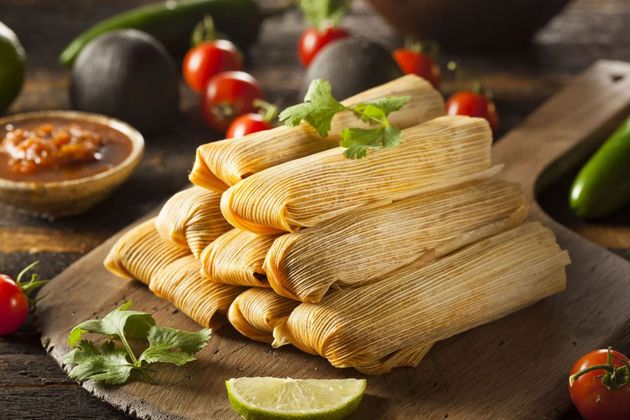 A stack of corn husk-wrapped tamales on a wooden cutting board with tomatoes, jalapeños, lime, and salsa nearby.