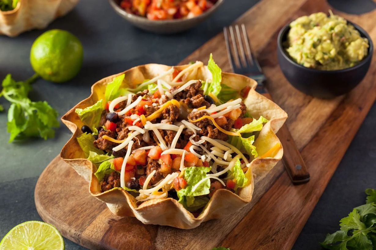 A taco salad in a crispy tortilla bowl, served on a wooden board with small bowls of guacamole and salsa nearby.