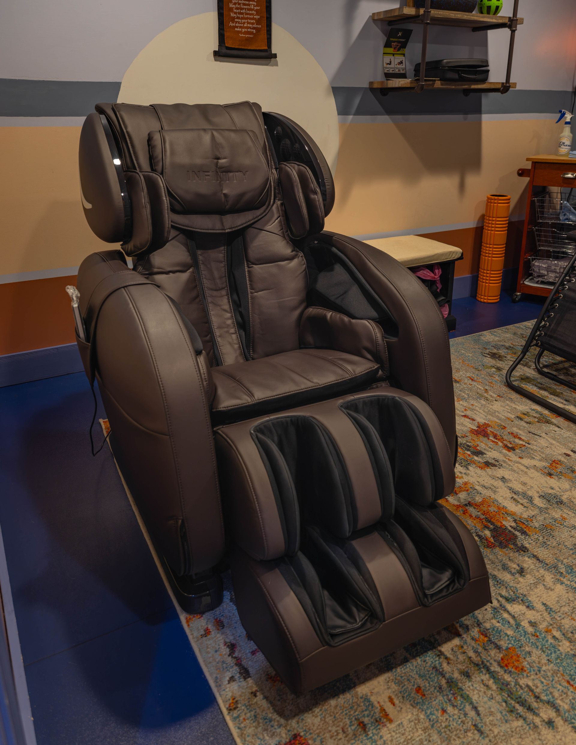 A dark brown, high-back massage chair sits on a textured rug in a room with a shelf and small table in the background.