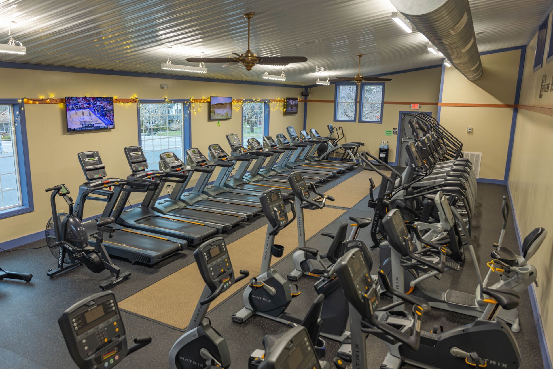 Rows of treadmills and stationary bikes line a gym with a textured ceiling, TVs, and large windows on a beige wall.
