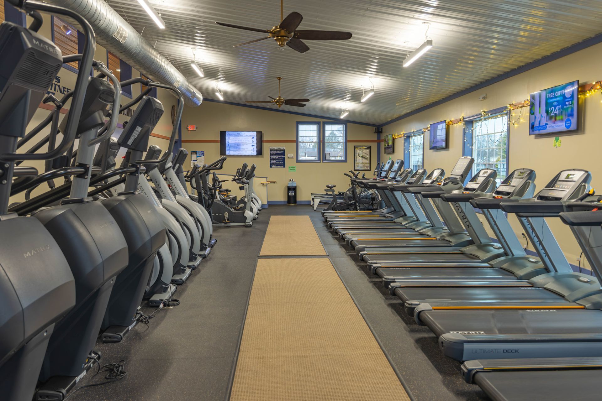 A bright, empty fitness center with rows of elliptical machines on the left and treadmills on the right.