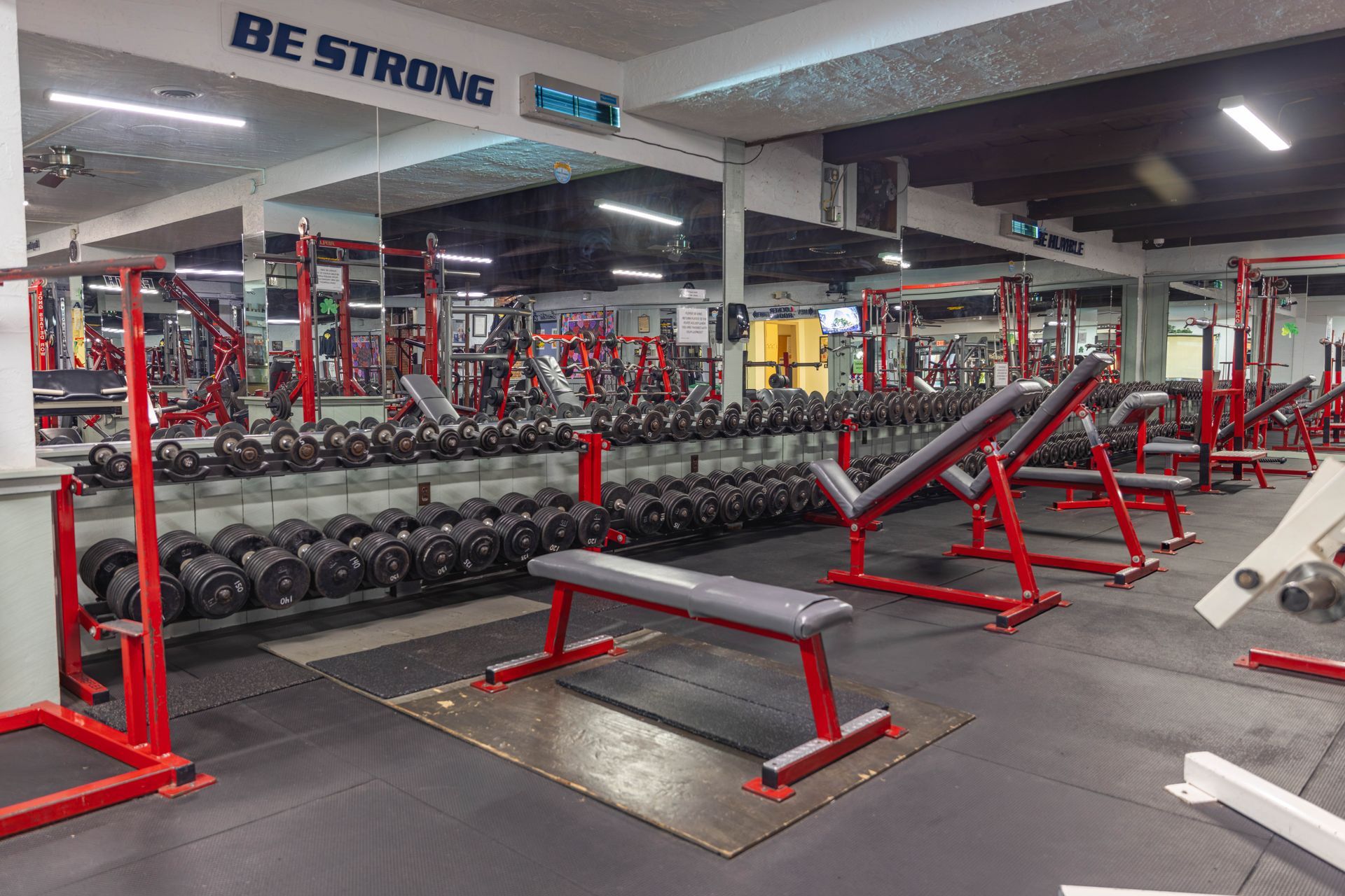 A gym featuring rows of black dumbbells on racks, a red weight bench, and a mirror wall with the text 