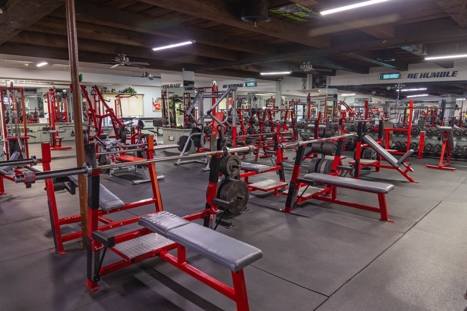 An empty gym featuring rows of red strength-training equipment, weight benches, and racks on a dark rubber mat floor.