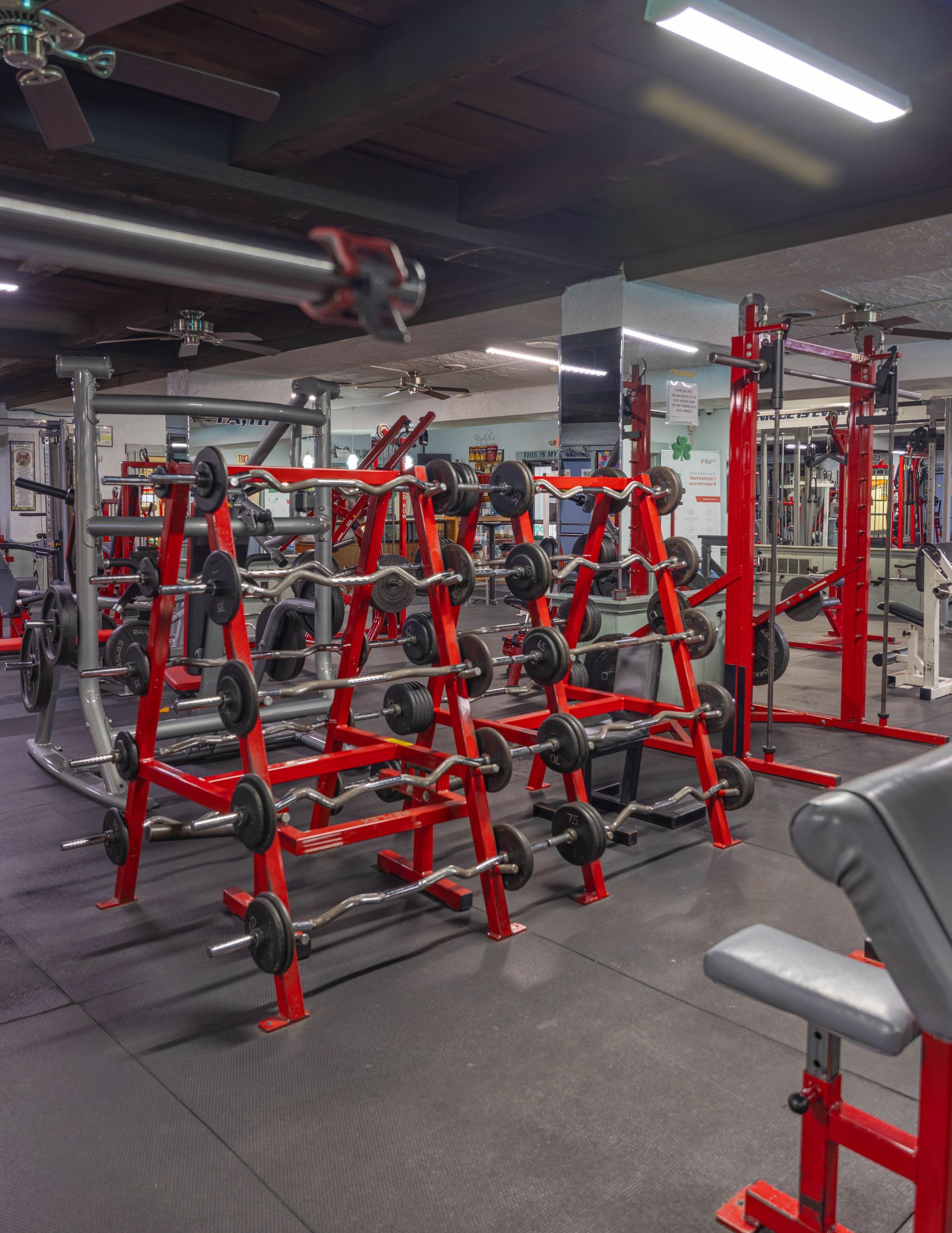 A gym interior features rows of red racks filled with various weight bars and plates under bright overhead lighting.