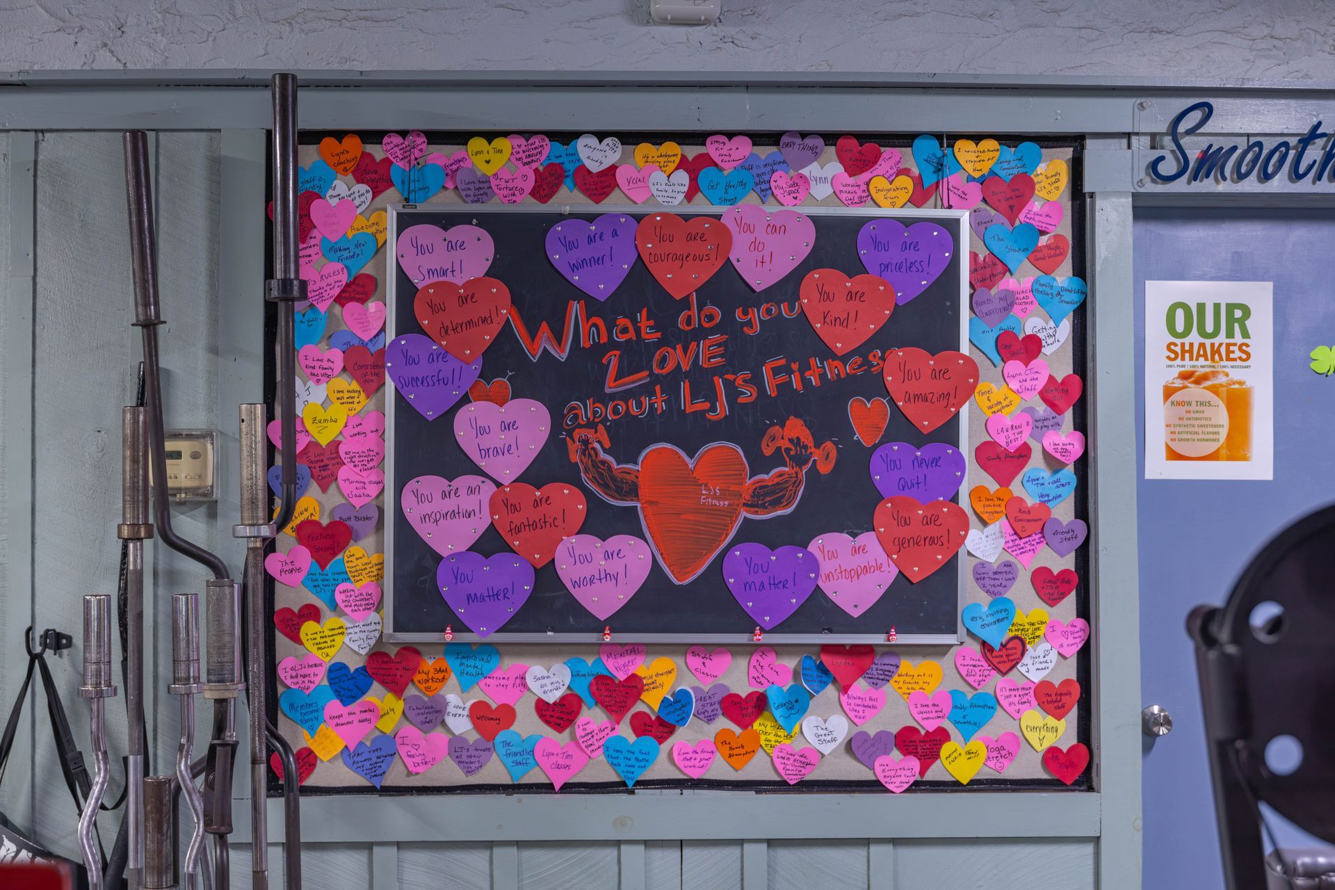 A gym bulletin board covered in colorful paper hearts with written messages surrounding a central heart drawing.