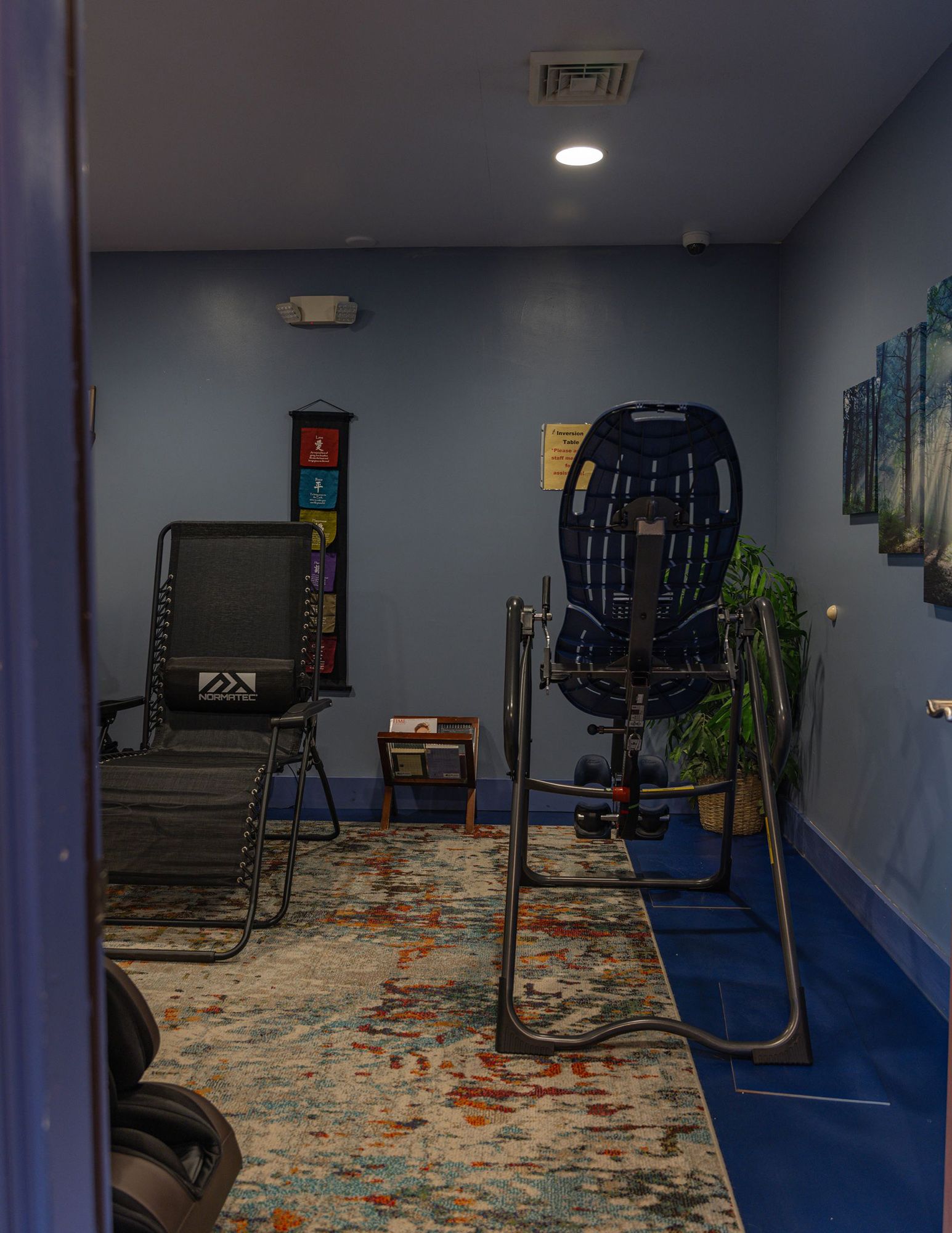 A blue-walled wellness room featuring an inversion table, a reclining chair, a massage chair, and a patterned area rug.