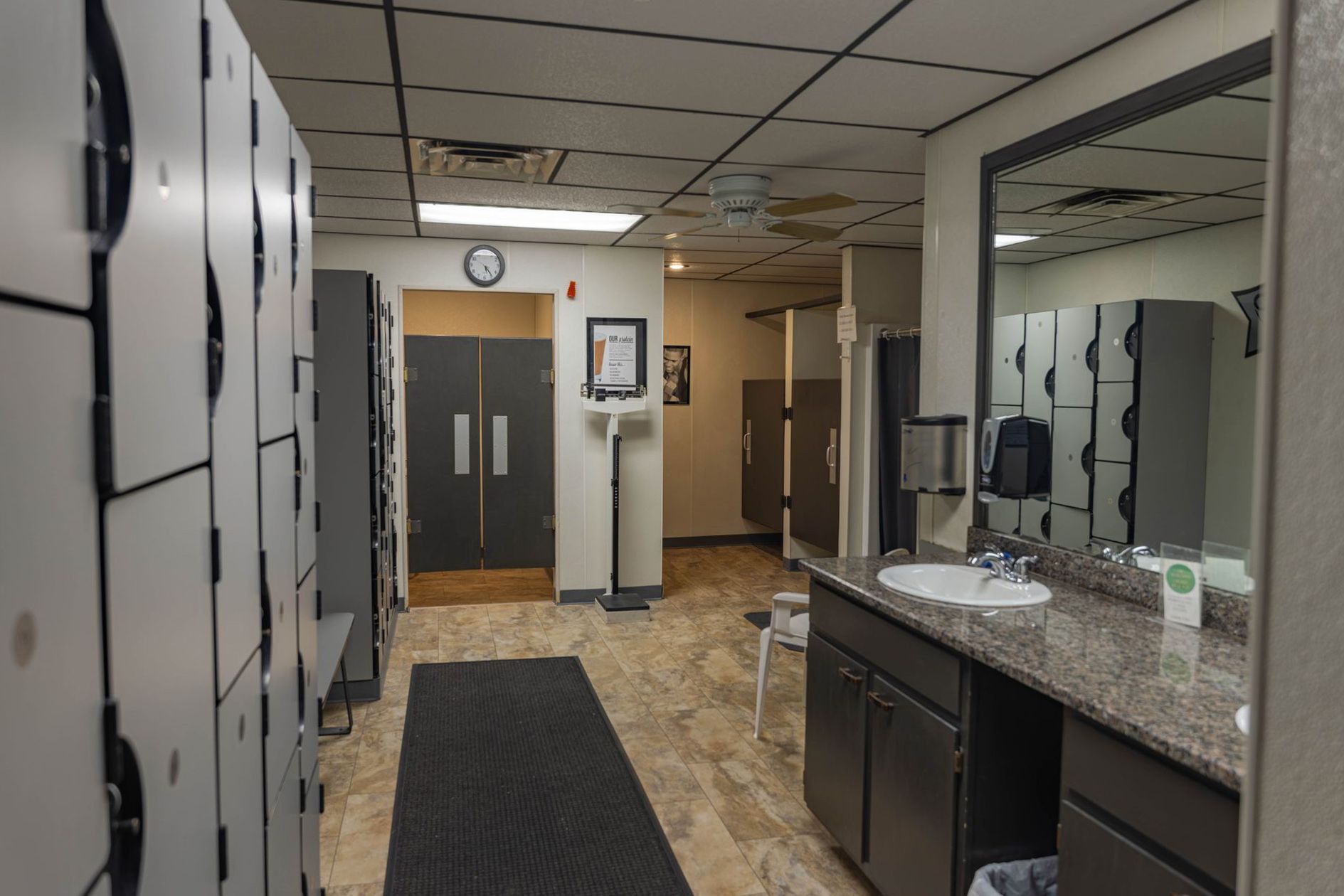 A locker room with rows of gray lockers on the left, a vanity with a sink on the right, and doors leading to a restroom.
