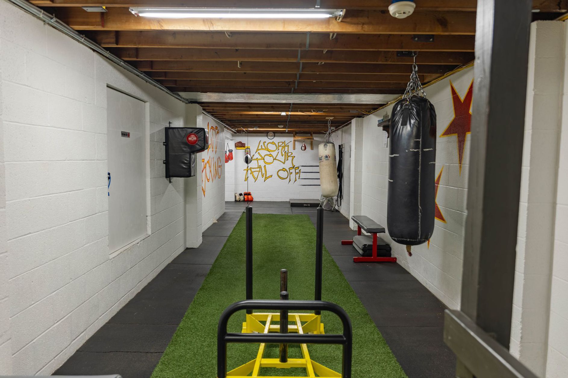 A home gym featuring a central artificial turf track with a yellow weight sled, a hanging punching bag, and wall art.