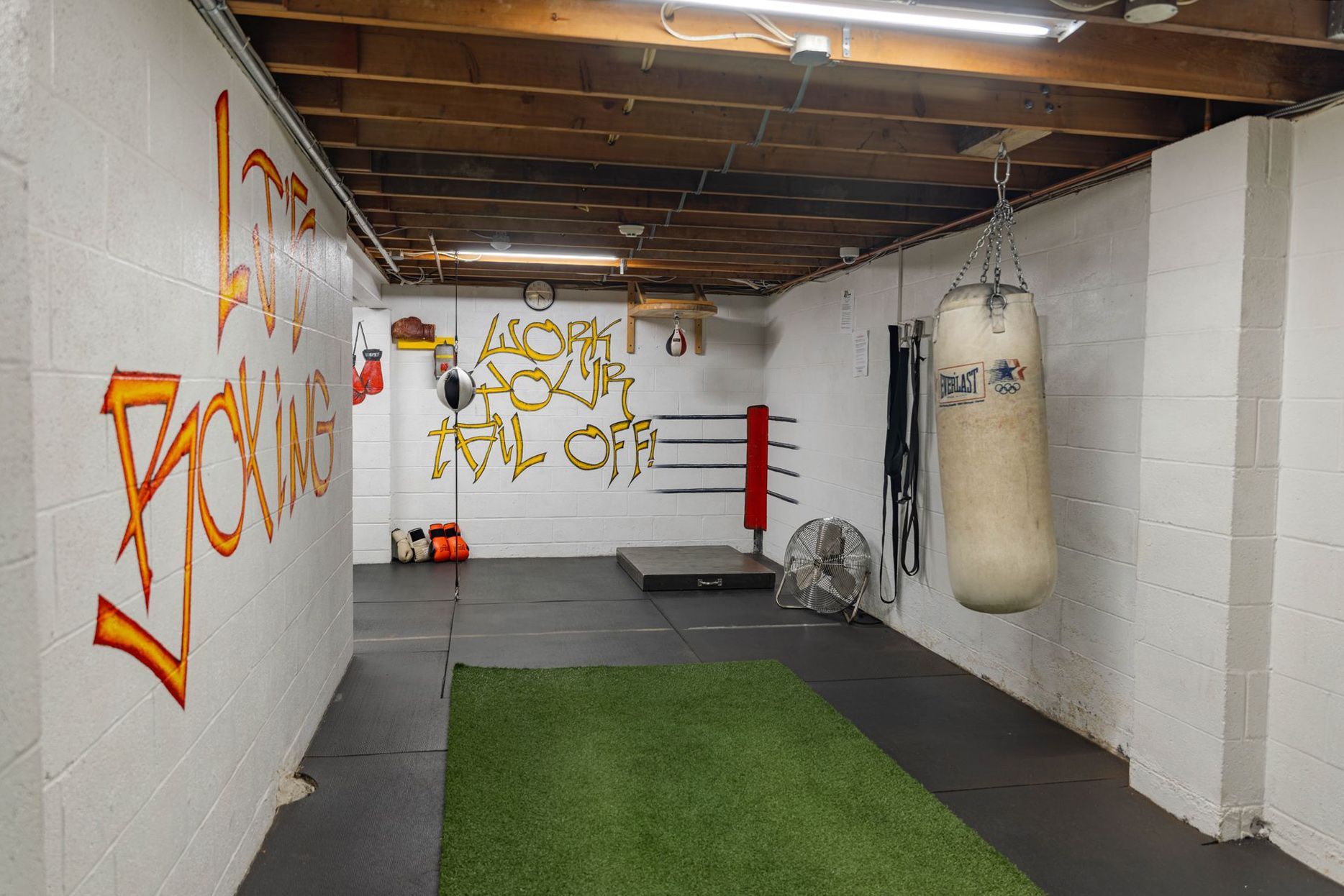A basement home gym featuring a punching bag, artificial turf floor, and graffiti art on white block walls.