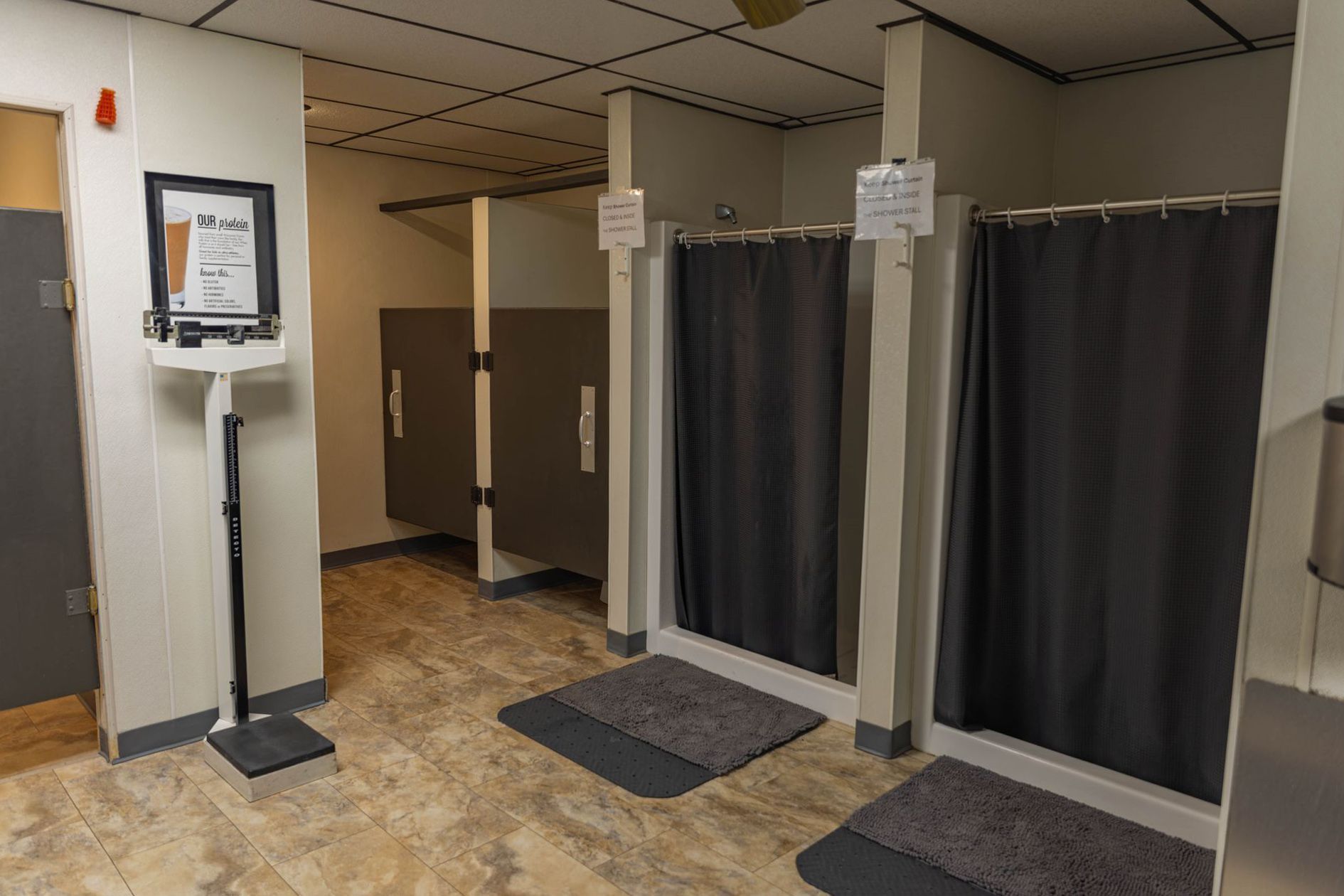 A public locker room area with a medical weight scale, privacy stalls, and two shower stalls with dark gray curtains.