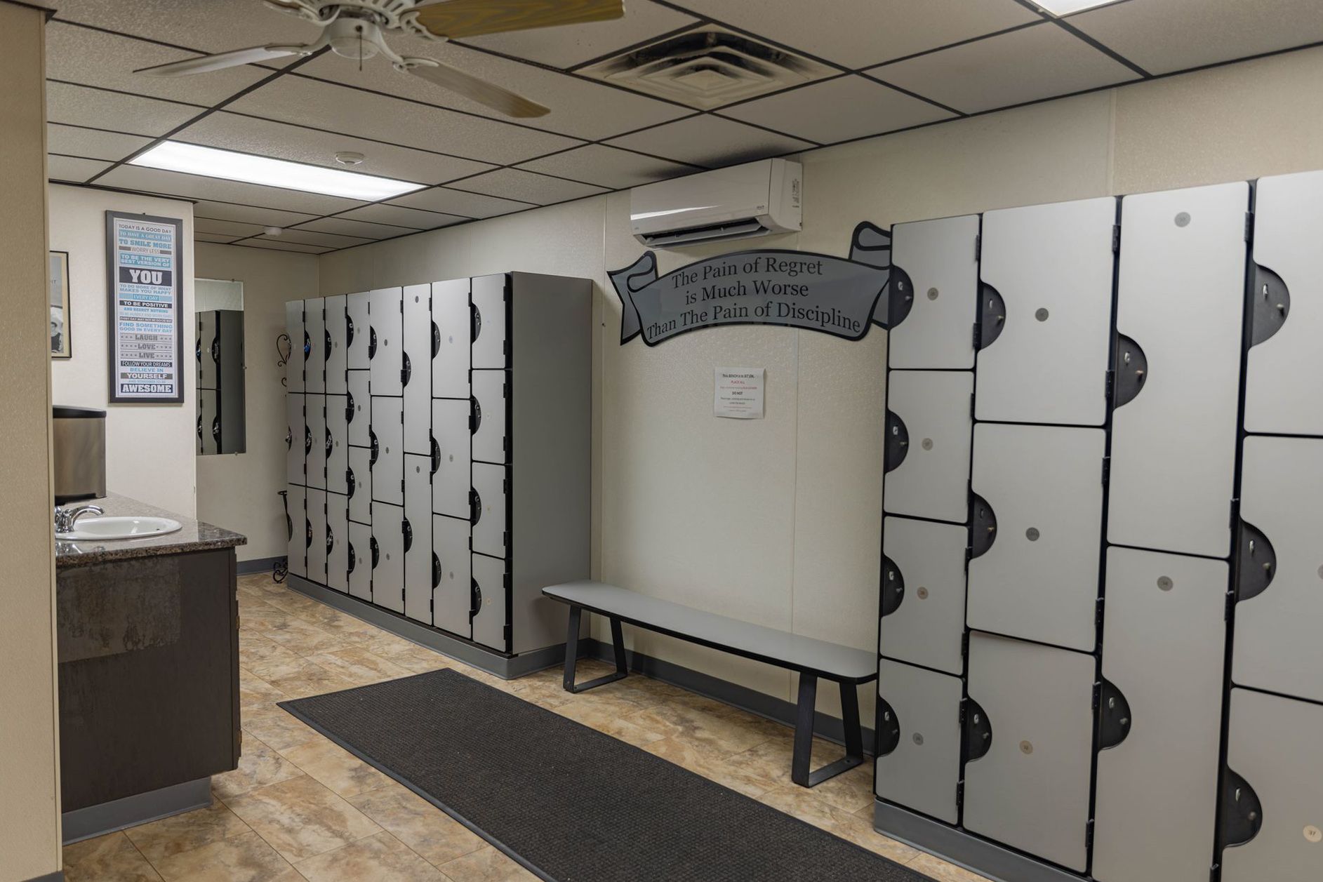 A locker room with gray lockers, a bench, a dark floor mat, and an air conditioning unit on a white wall.