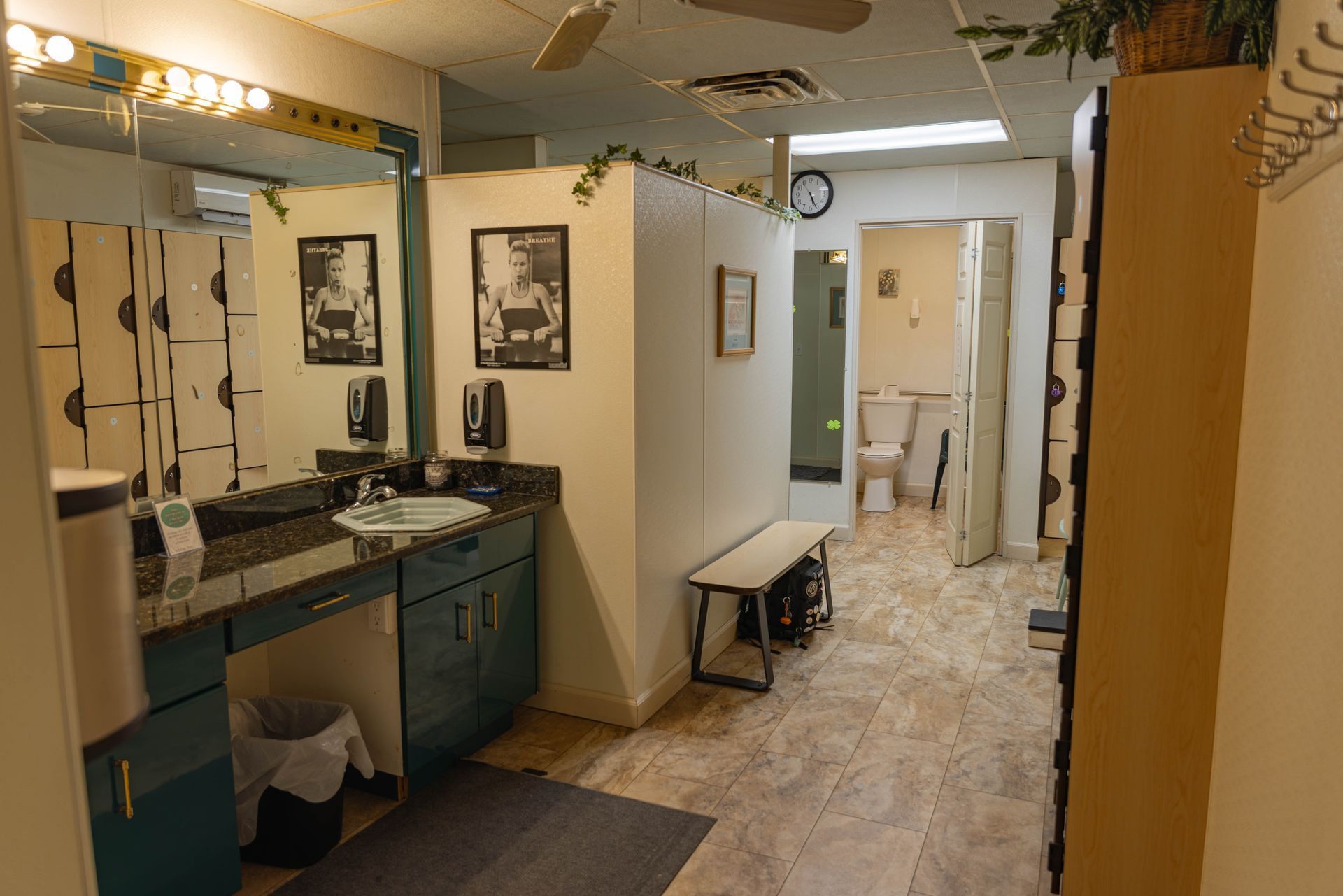 A brightly lit, tiled restroom featuring a dark blue vanity with a sink, a bench, and an open doorway to a toilet.
