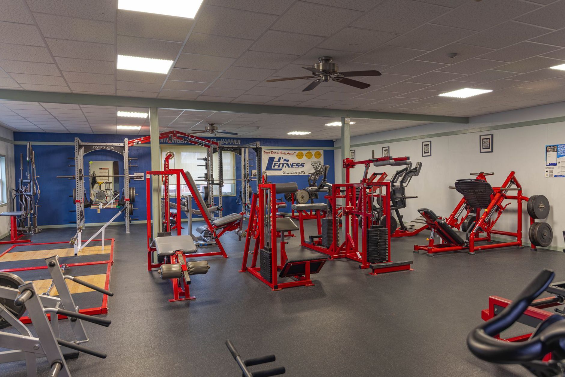 An empty gym interior featuring several red strength training machines, power racks, and floor mats.