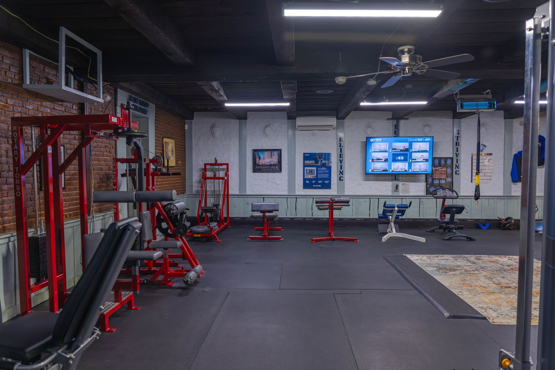 An indoor gym with red weight equipment, gym flooring, and a large flat-screen display on a white wall.
