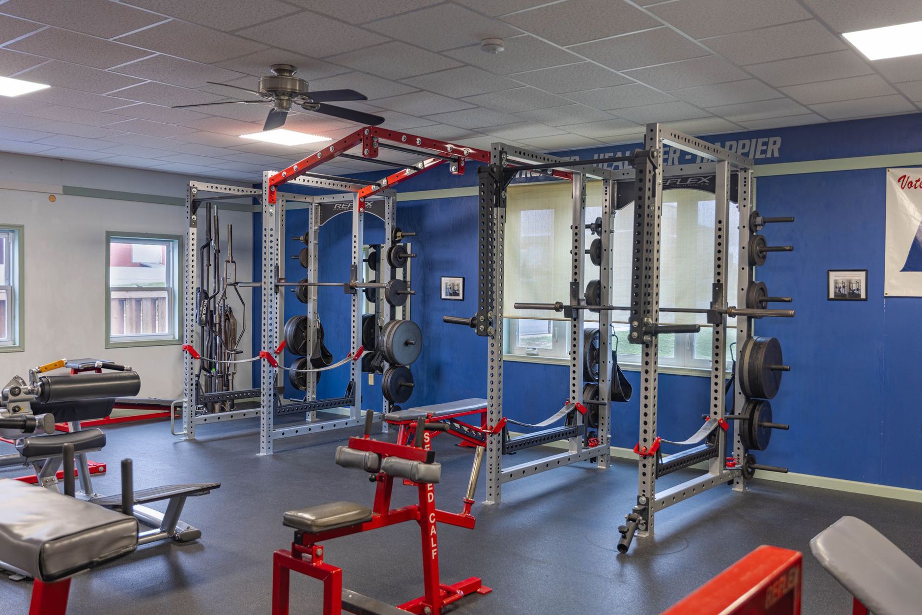A gym interior with empty weight racks, weight plates, and benches against a blue wall.