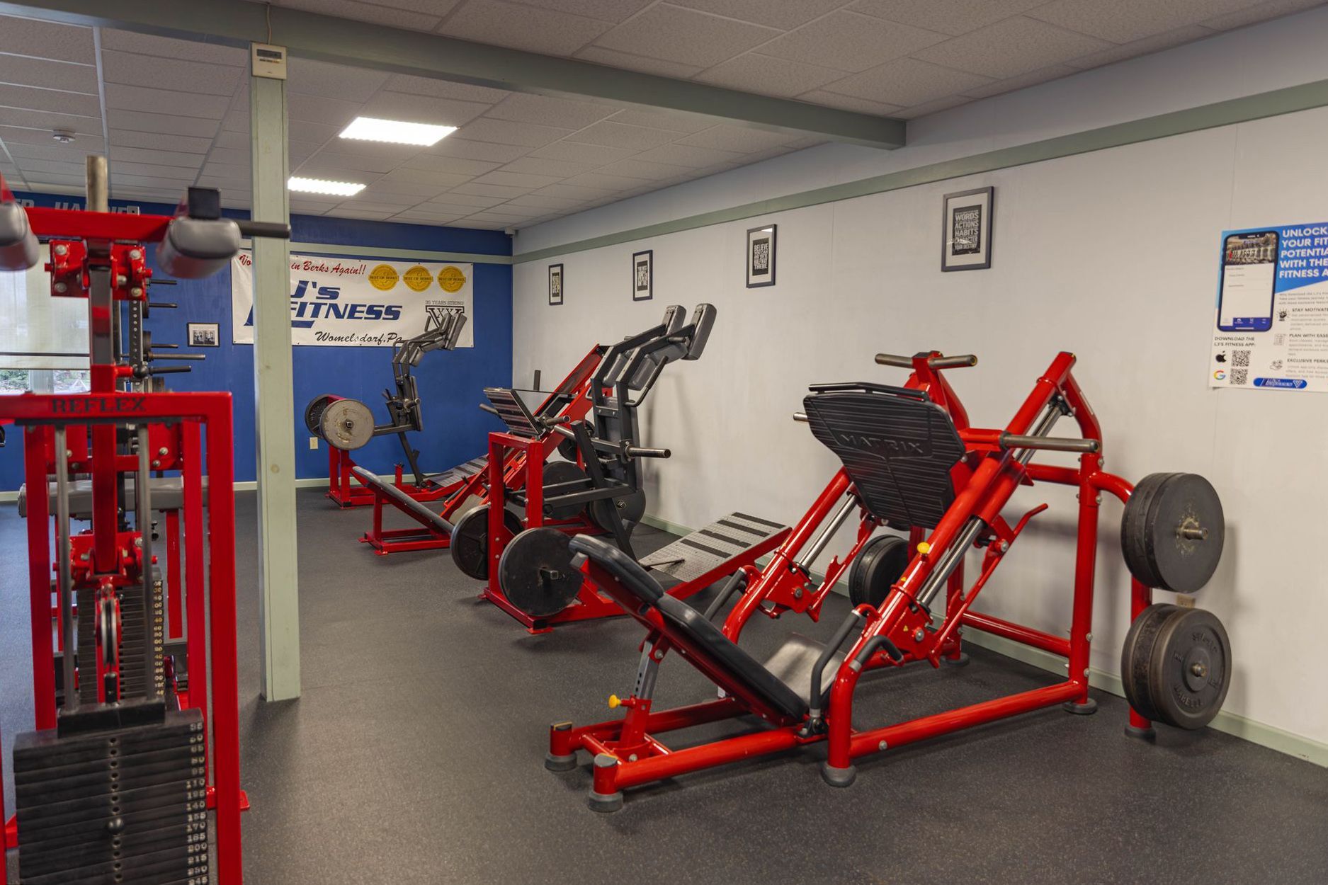 A gym interior with several red strength training machines, including a leg press, arranged in a row.
