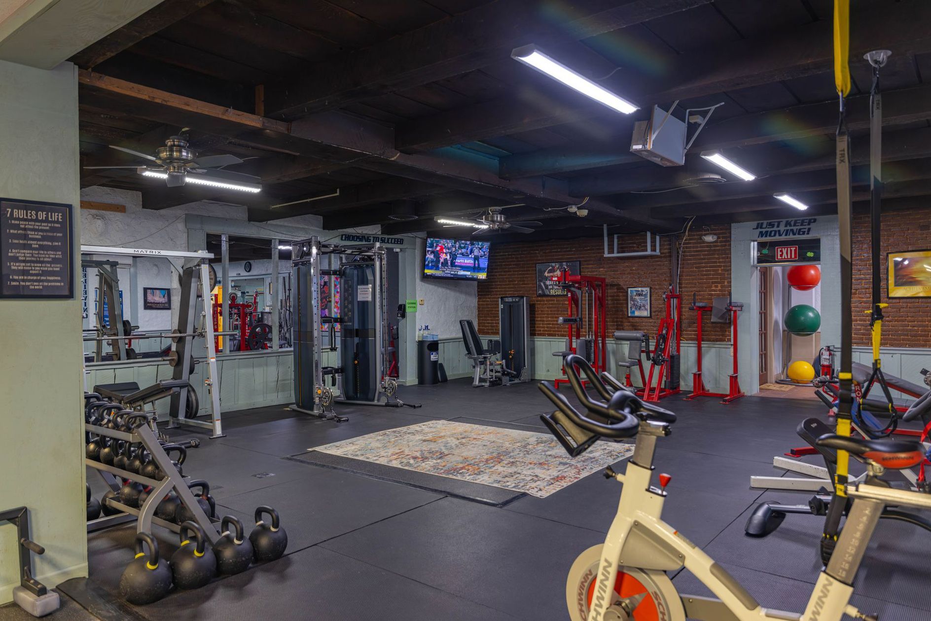 A basement gym with rubber flooring, a stationary bike in the foreground, weight racks, kettlebells, and a flat-screen TV.
