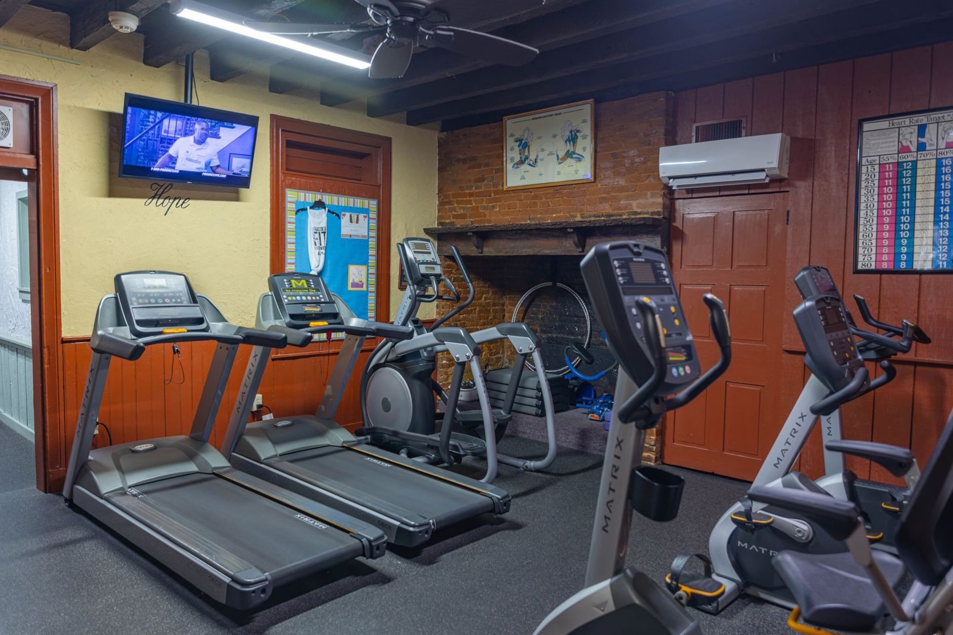 A small, dimly lit home gym with two treadmills, a stationary bike, and an elliptical machine against a brick wall.