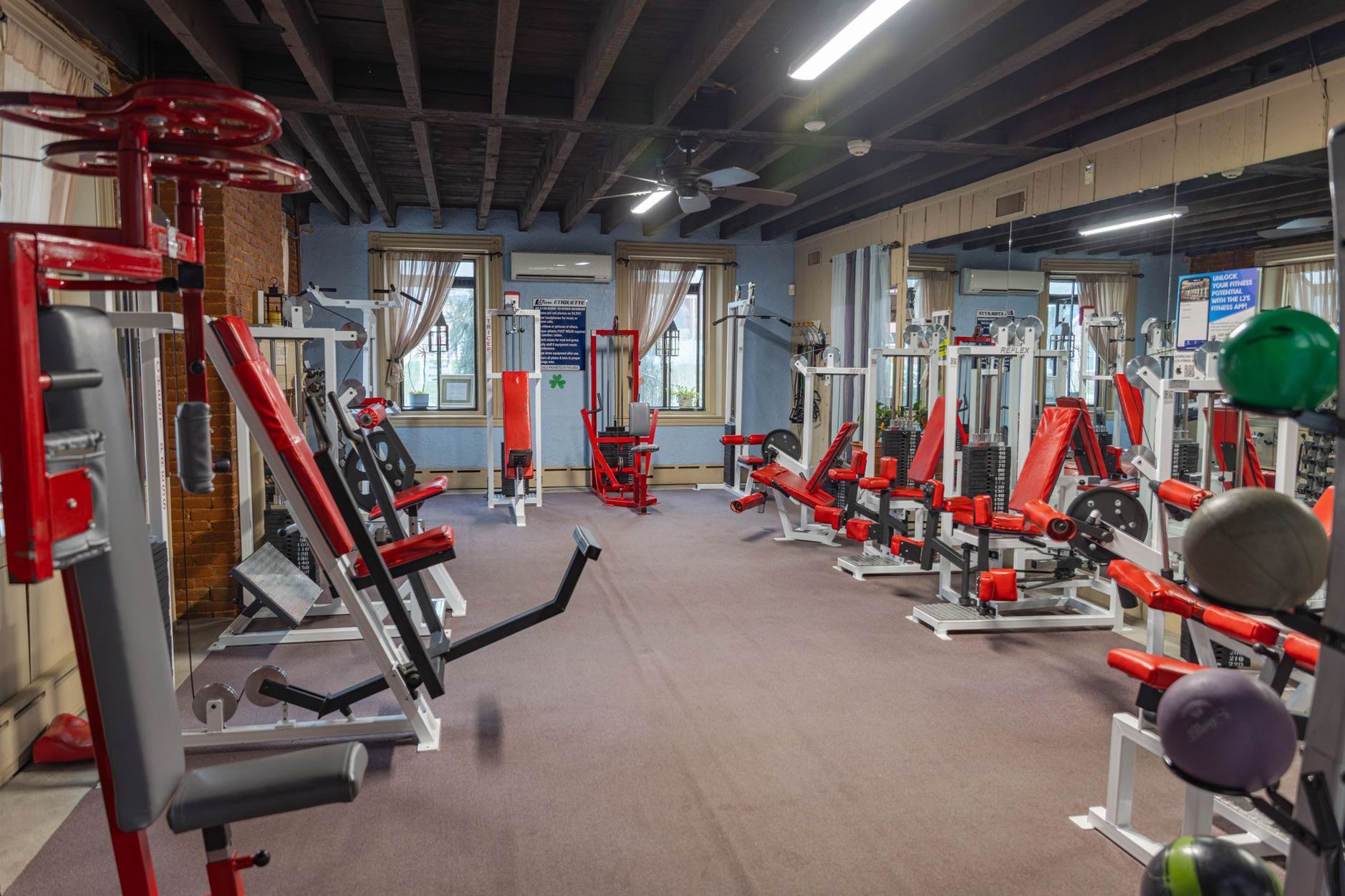Indoor gym with rows of red exercise machines, a carpeted floor, and windows on a back wall.