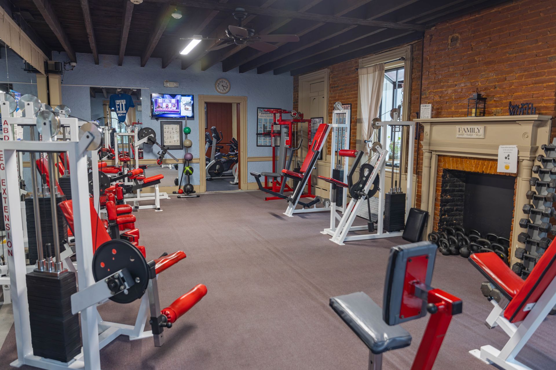 A home gym with red weight machines, free weights, and a brick fireplace, set in a room with a dark wood-beamed ceiling.