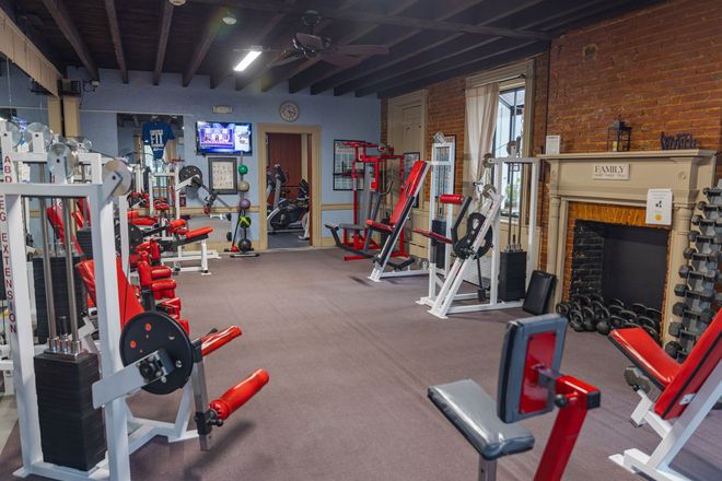 A home gym with red weight machines, free weights, and a brick fireplace, set in a room with a dark wood-beamed ceiling.