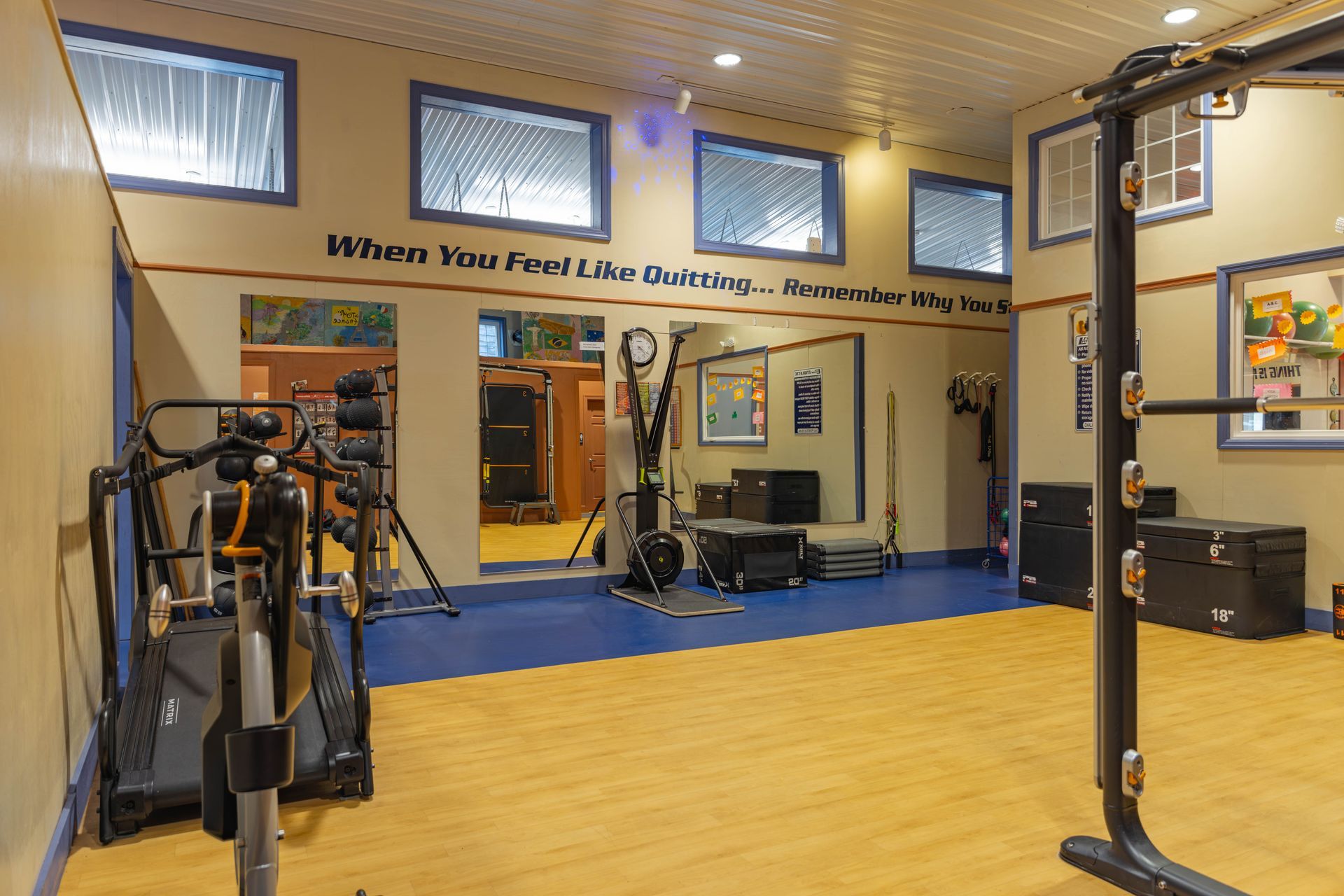 A home gym with gym equipment, blue and wood flooring, windows, and an inspirational quote painted on the wall.