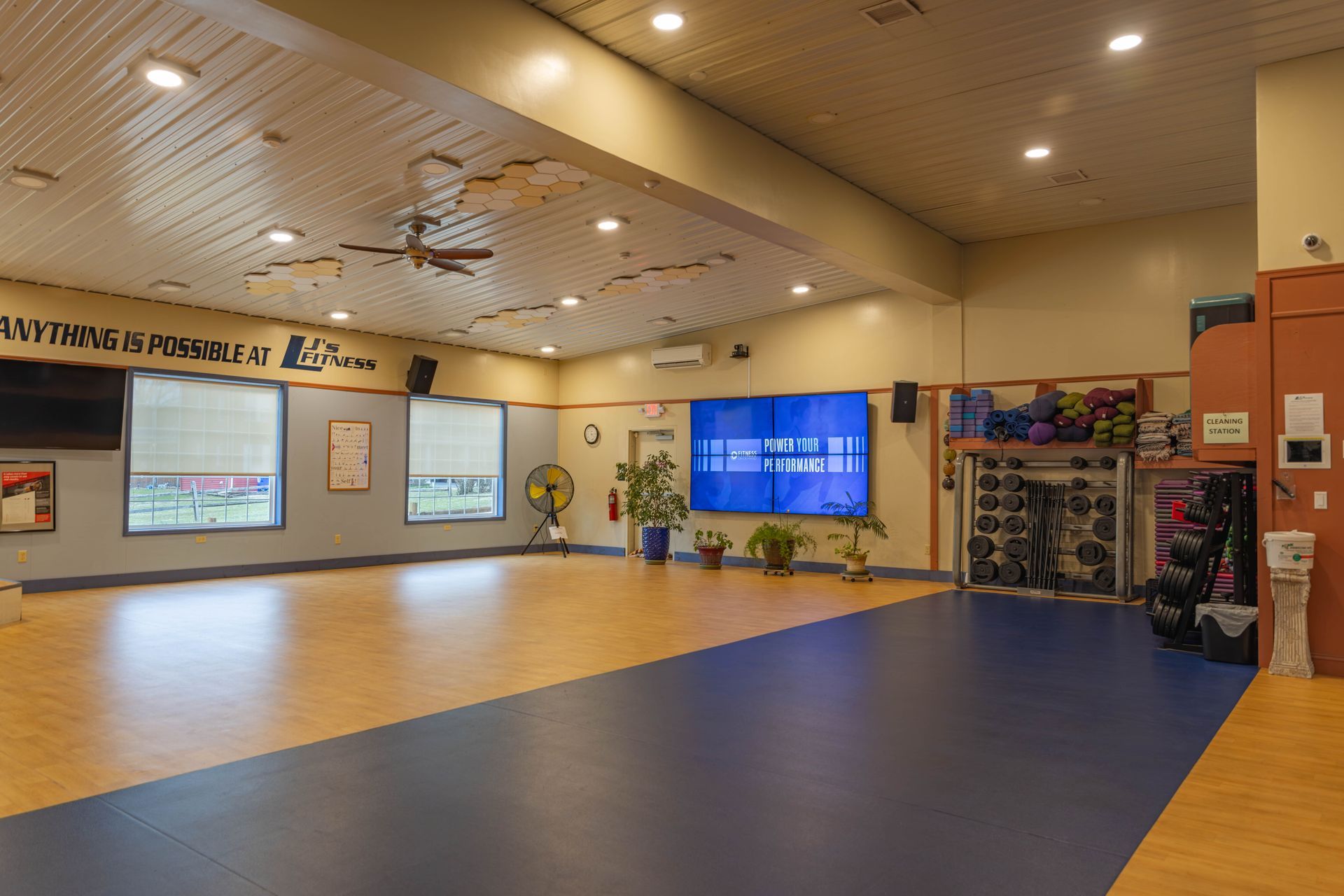 A spacious fitness studio with wood and blue flooring, ceiling fans, large windows, and storage racks for equipment.