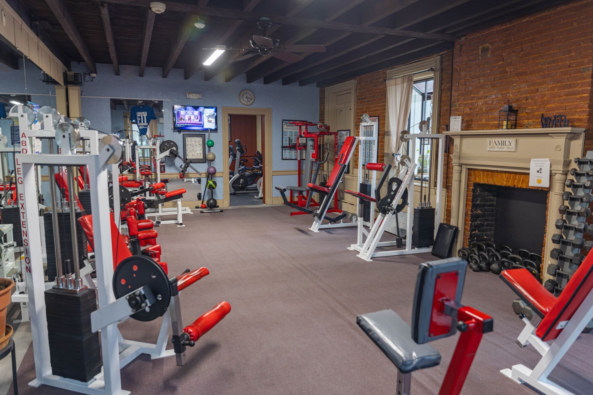 A gym with various weight machines, a rack of dumbbells, and a fireplace, set in a room with dark ceiling beams.