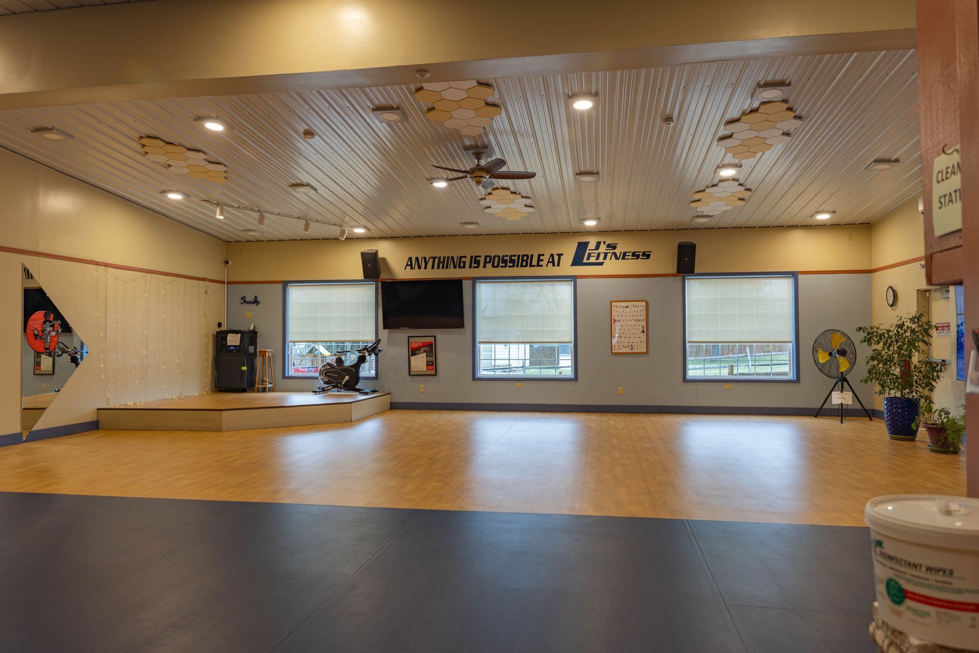 A spacious indoor dance studio with wooden floors, a small stage area, neutral-colored walls, and recessed ceiling lights.