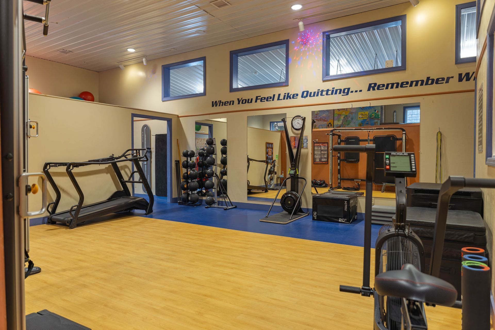 A home gym with light wood floors, a treadmill, medicine balls, and fitness equipment against blue and tan walls.