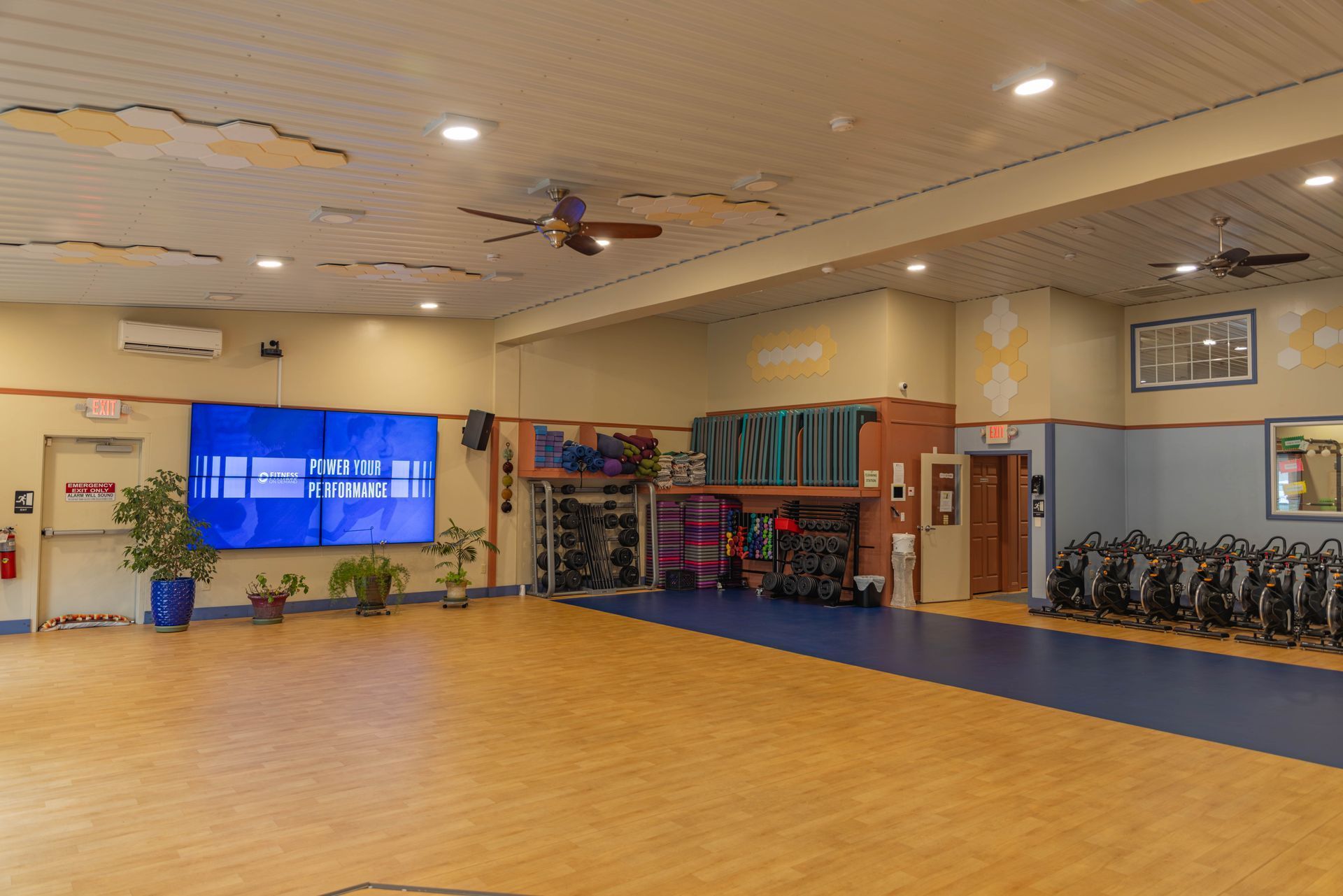 A wide, empty fitness studio with wooden flooring, exercise bikes, shelving for mats, and a large blue screen on the wall.