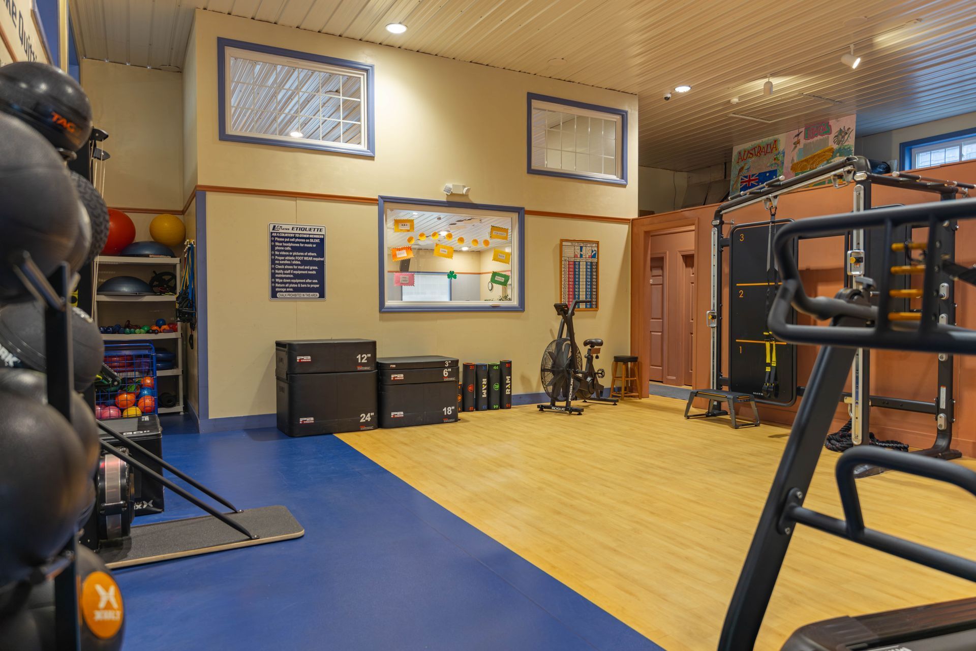 A well-lit, organized fitness gym with wood flooring, blue mats, weight equipment, and a stationary exercise bike.