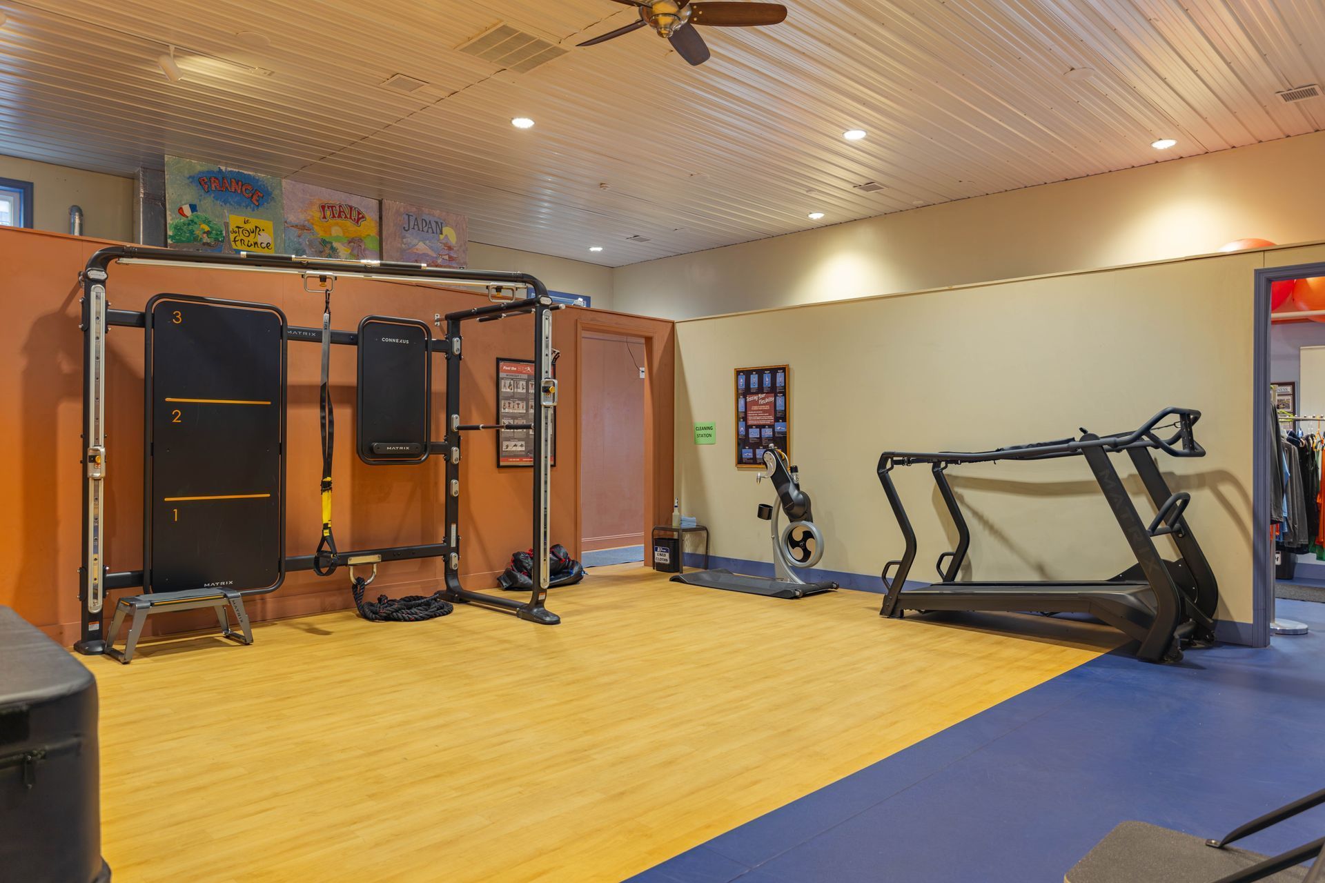 A gym interior with a workout rig, a treadmill, and a stationary bike on a wooden floor, with tan walls and a blue area.