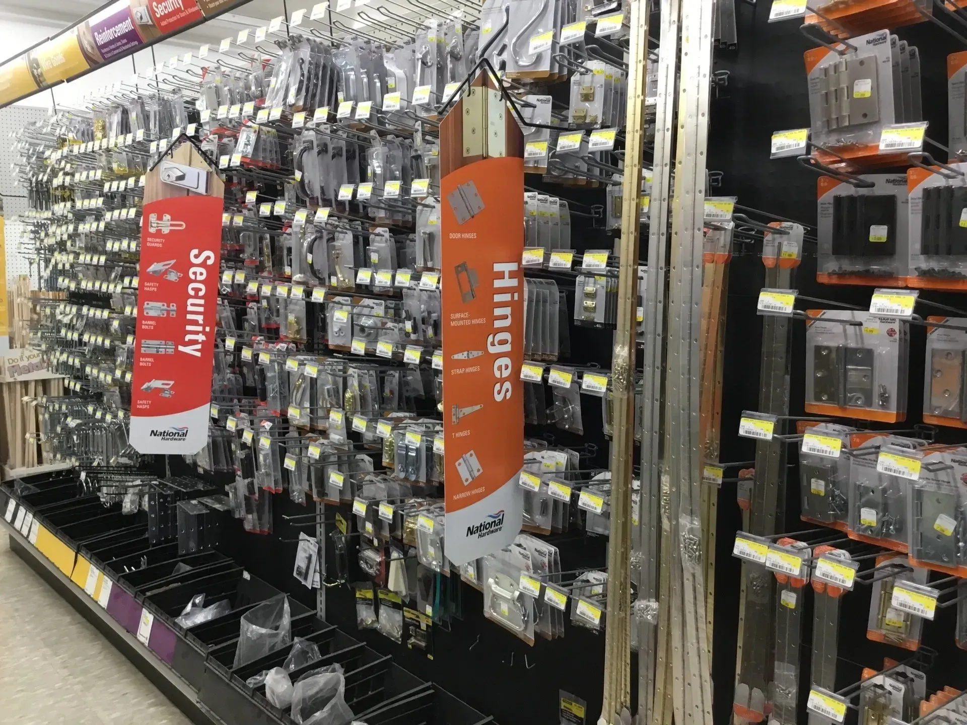 Hardware store aisle with hanging tools and accessories, marked by red “Security” and orange “Hinges” signs.