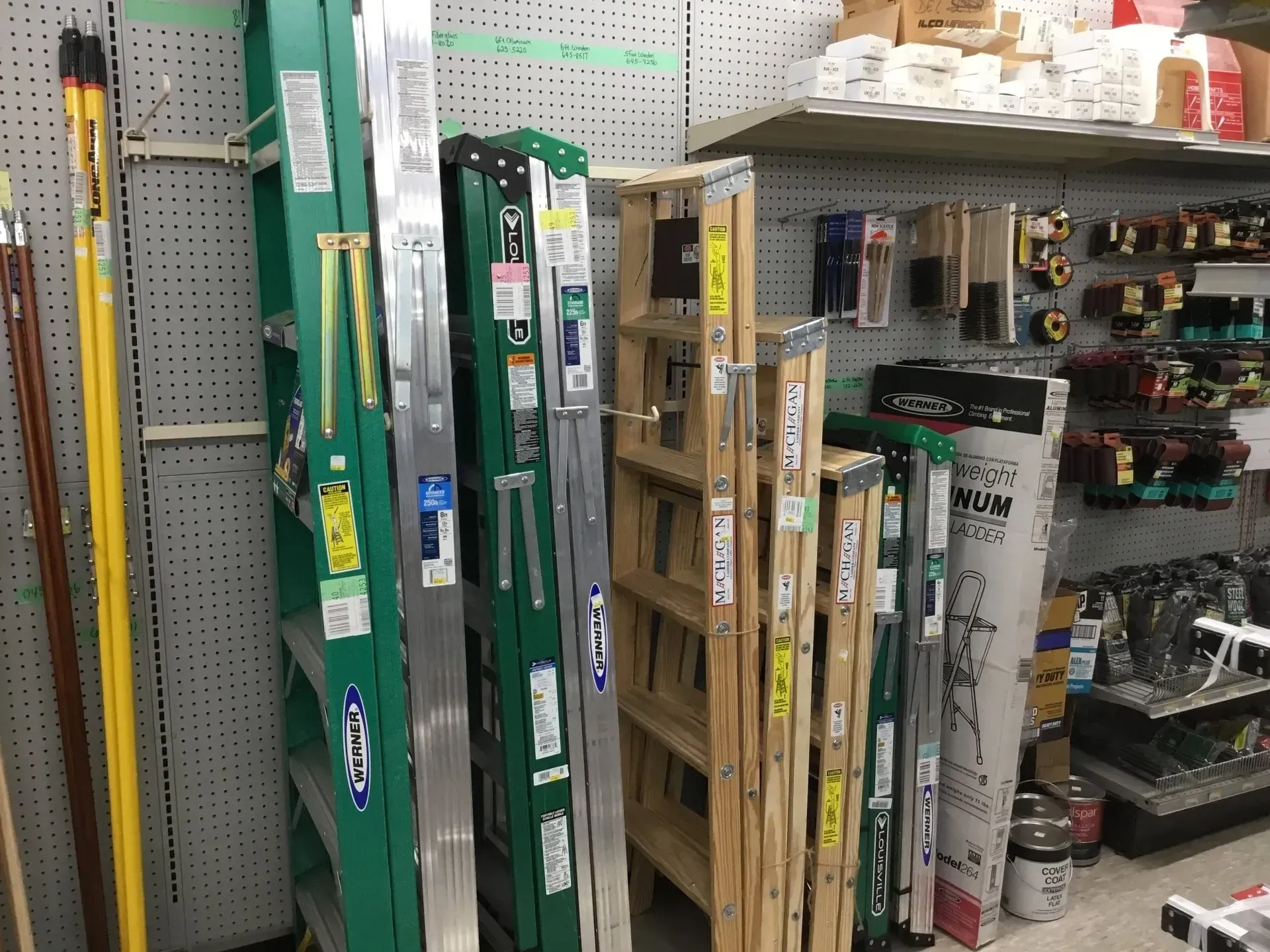 Hardware store aisle with long-handled tools in green packaging on shelves and a pegboard wall.