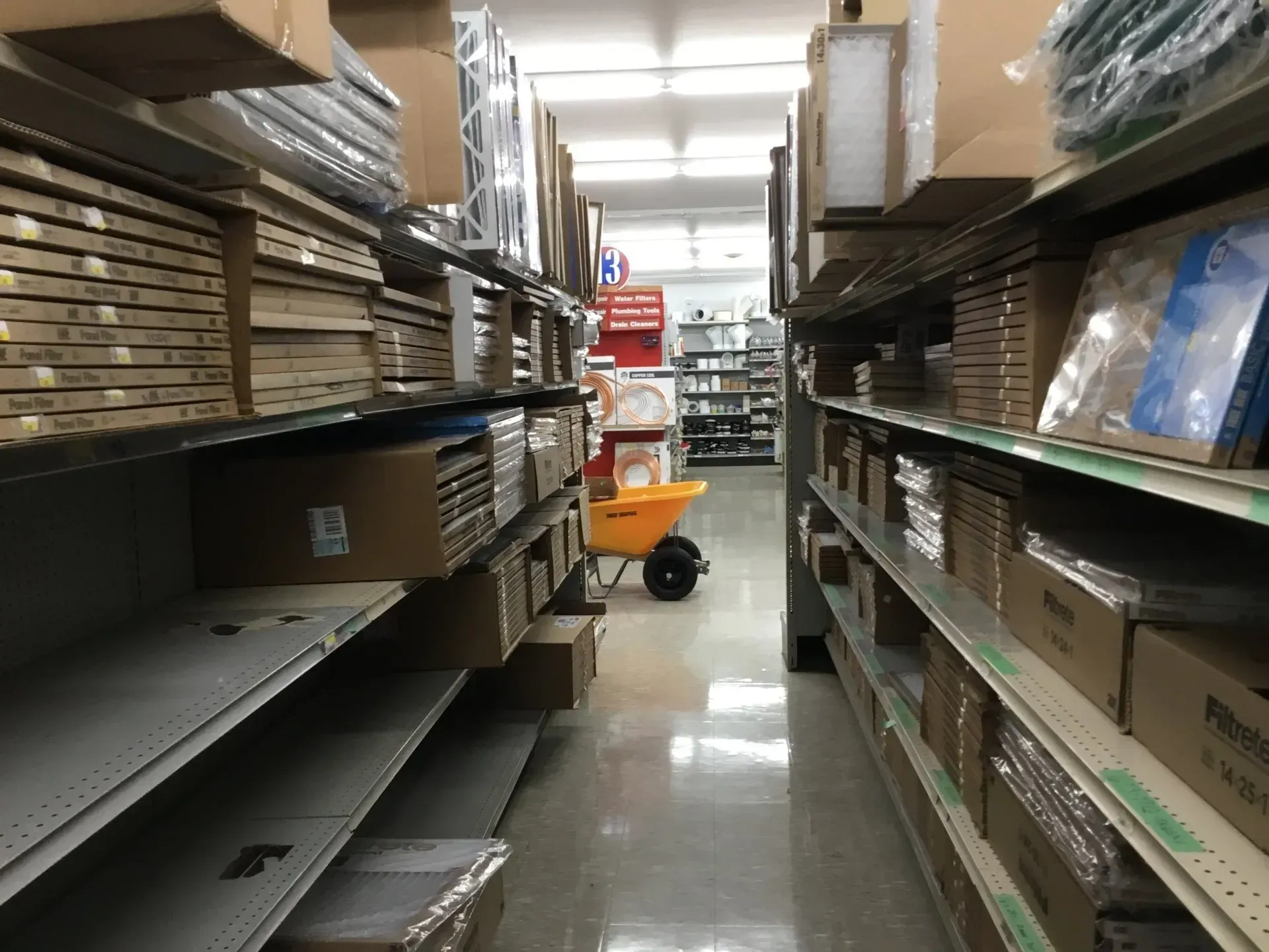 Aisle of a hardware store with shelves of boxes and supplies, plus a small orange cart in the distance.