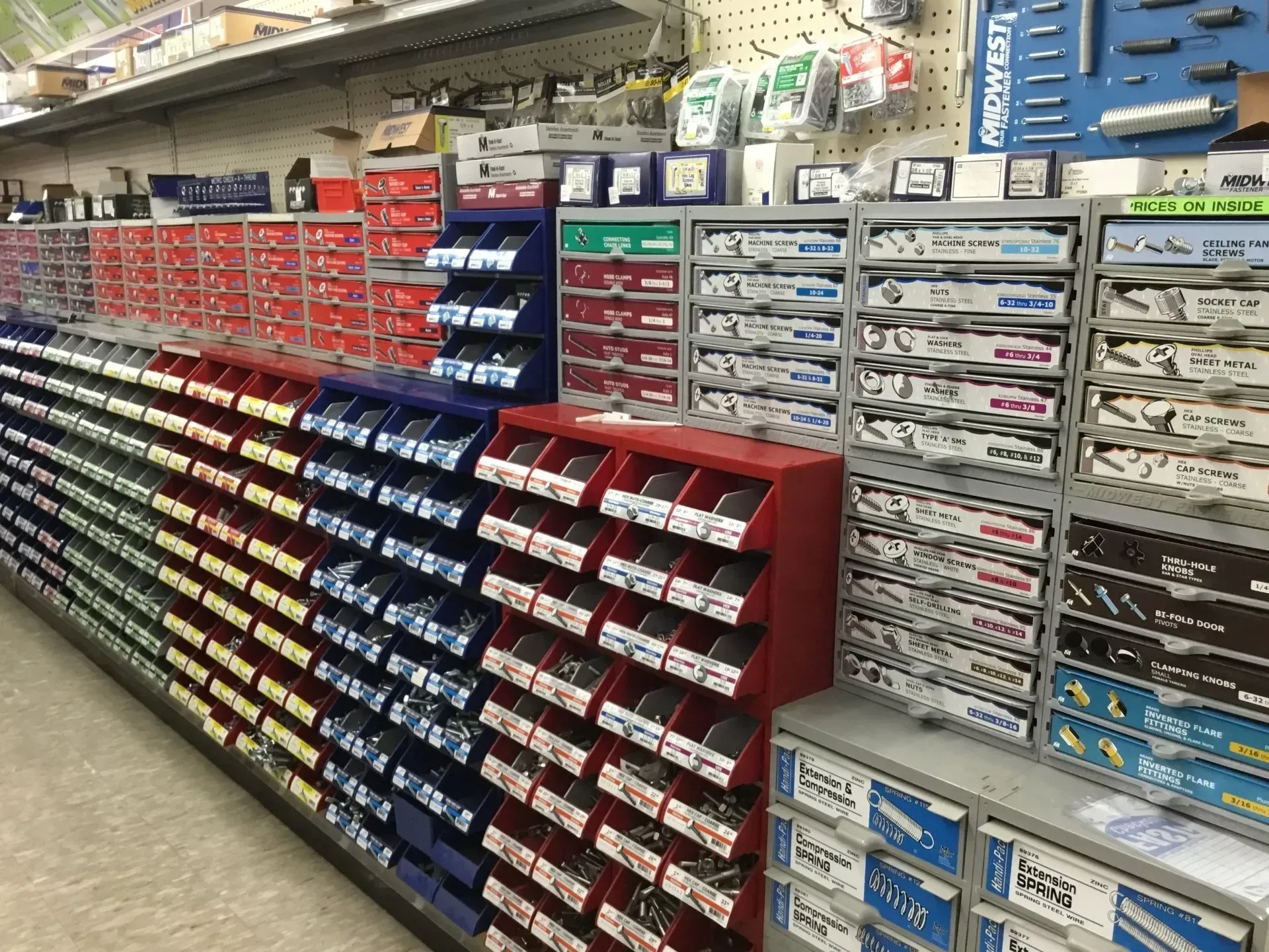 Aisle of hardware store shelves stocked with rows of small parts in red, blue, and gray bins
