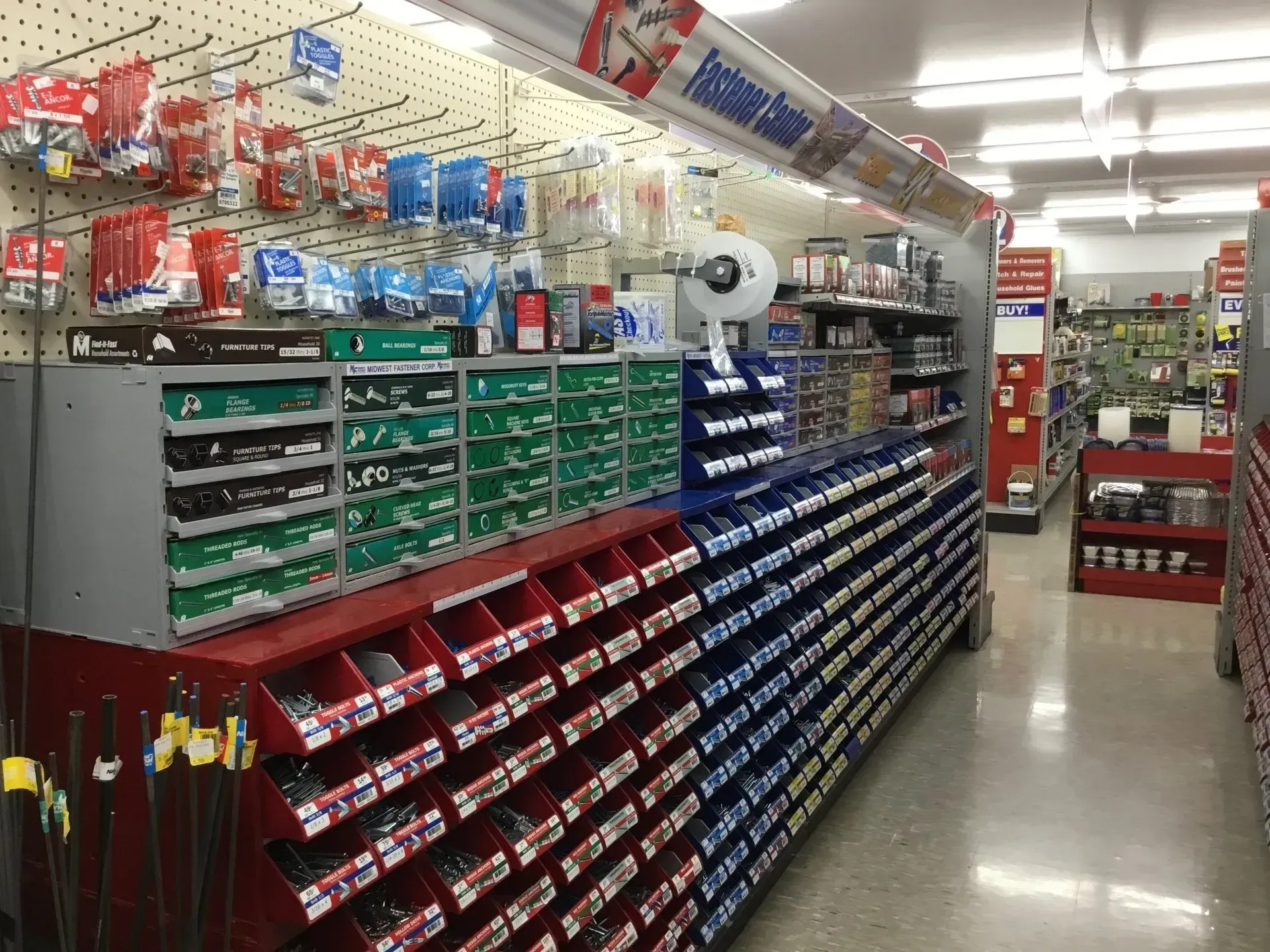 Store aisle with rows of hardware bins and supplies on shelves under bright fluorescent lights