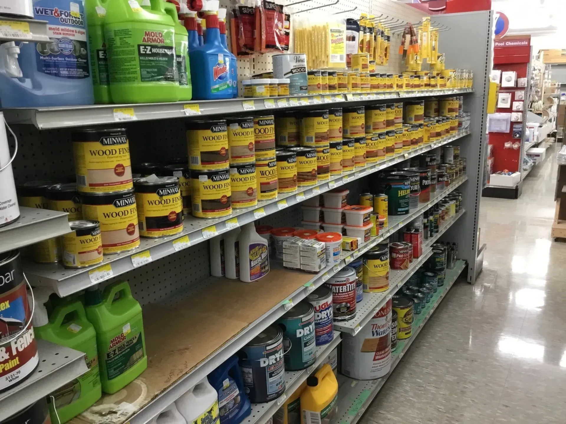 A store aisle stocked with paint cans and supplies on metal shelves, viewed from the side.