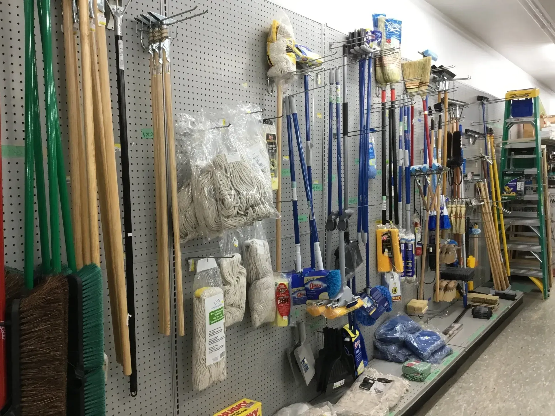 Assorted cleaning tools and mops hanging on a store pegboard aisle, with buckets and supplies below