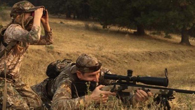 Two people hunting, one looking through binoculars, the other aiming a rifle with a scope, in a field