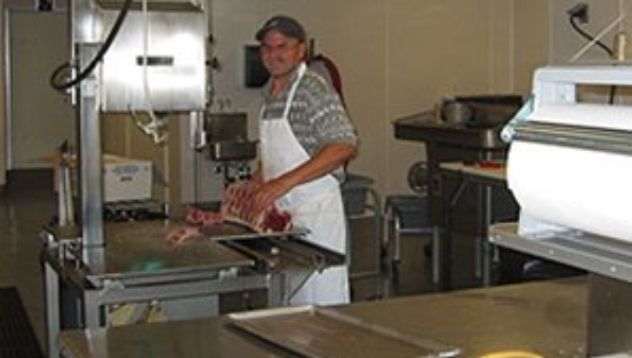 Butcher in a commercial kitchen using a band saw to cut meat, wearing an apron