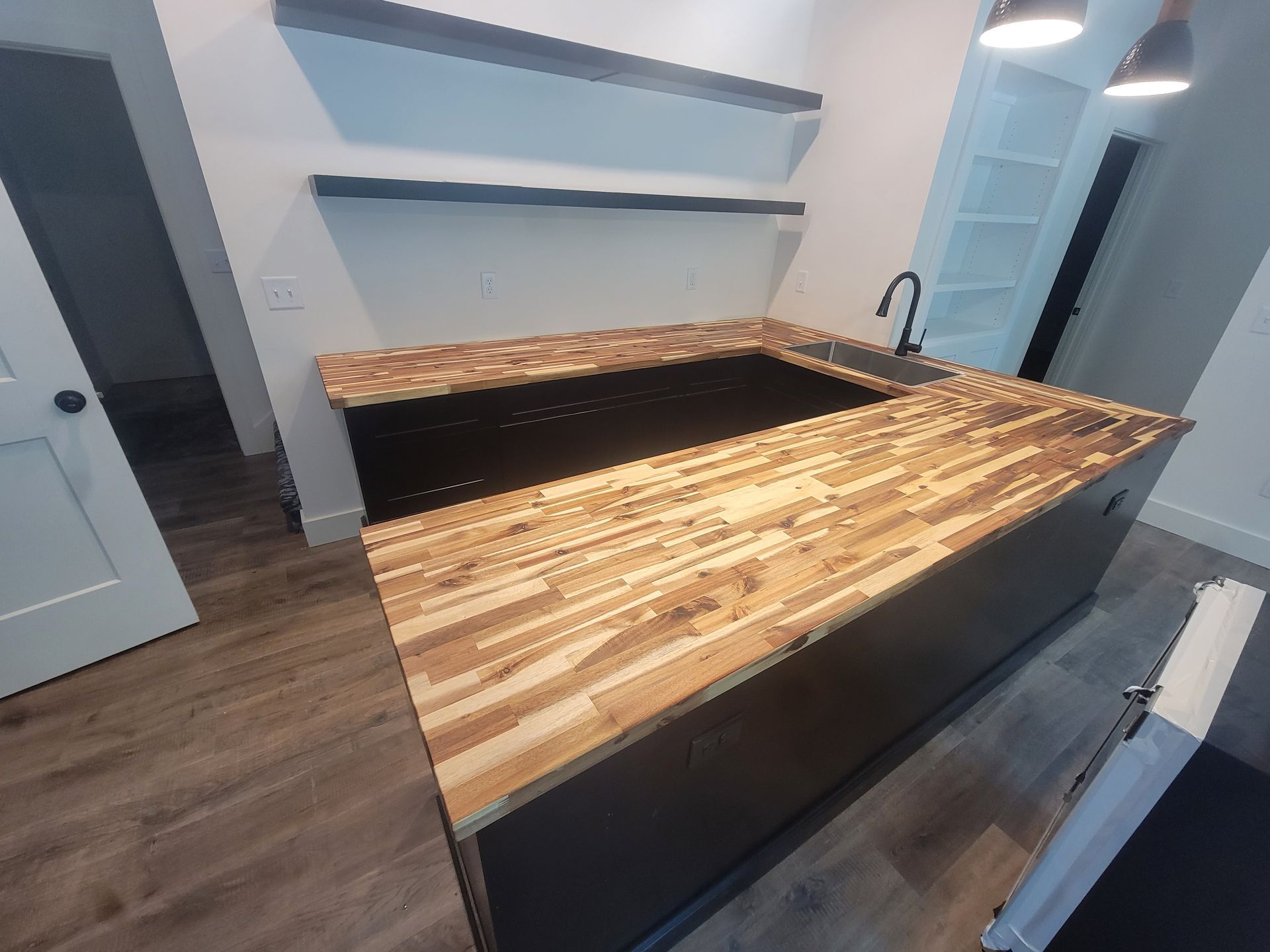Kitchen island with wood countertop and black cabinets, shelves on wall, sink.