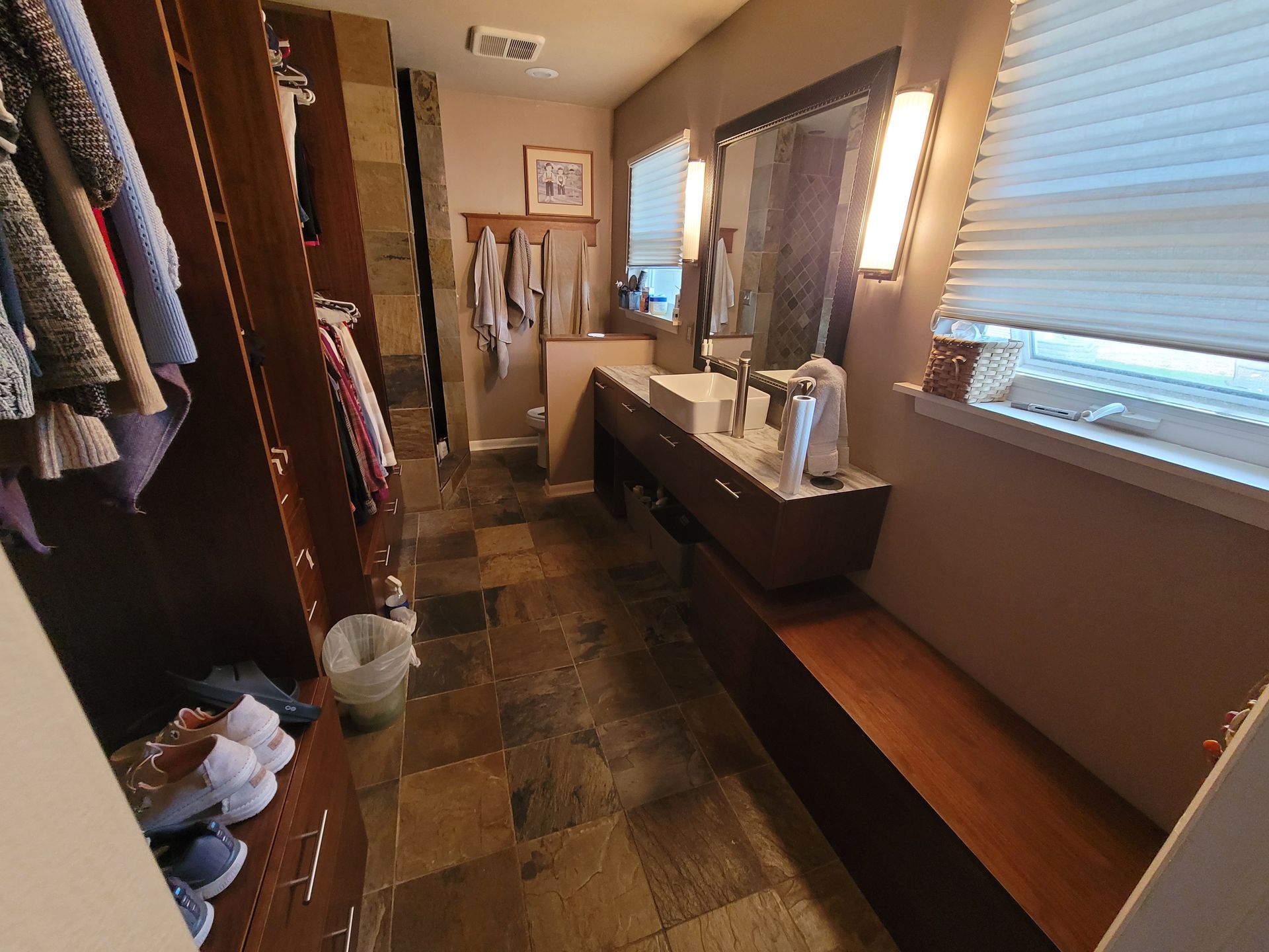 Walk-in closet with bathroom. Dark wood cabinets, stone tile floor. Sink, mirror, towels, and window with blinds.